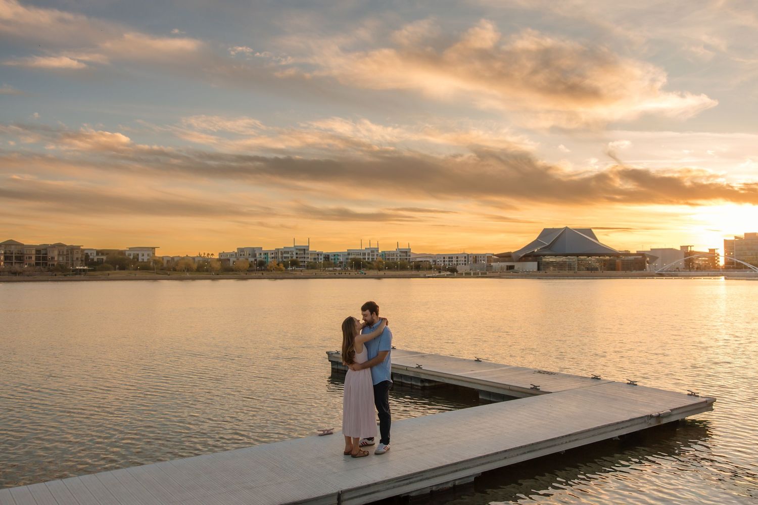 Romantic couple sharing a kiss on a dock at sunset over a calm lake with city skyline.