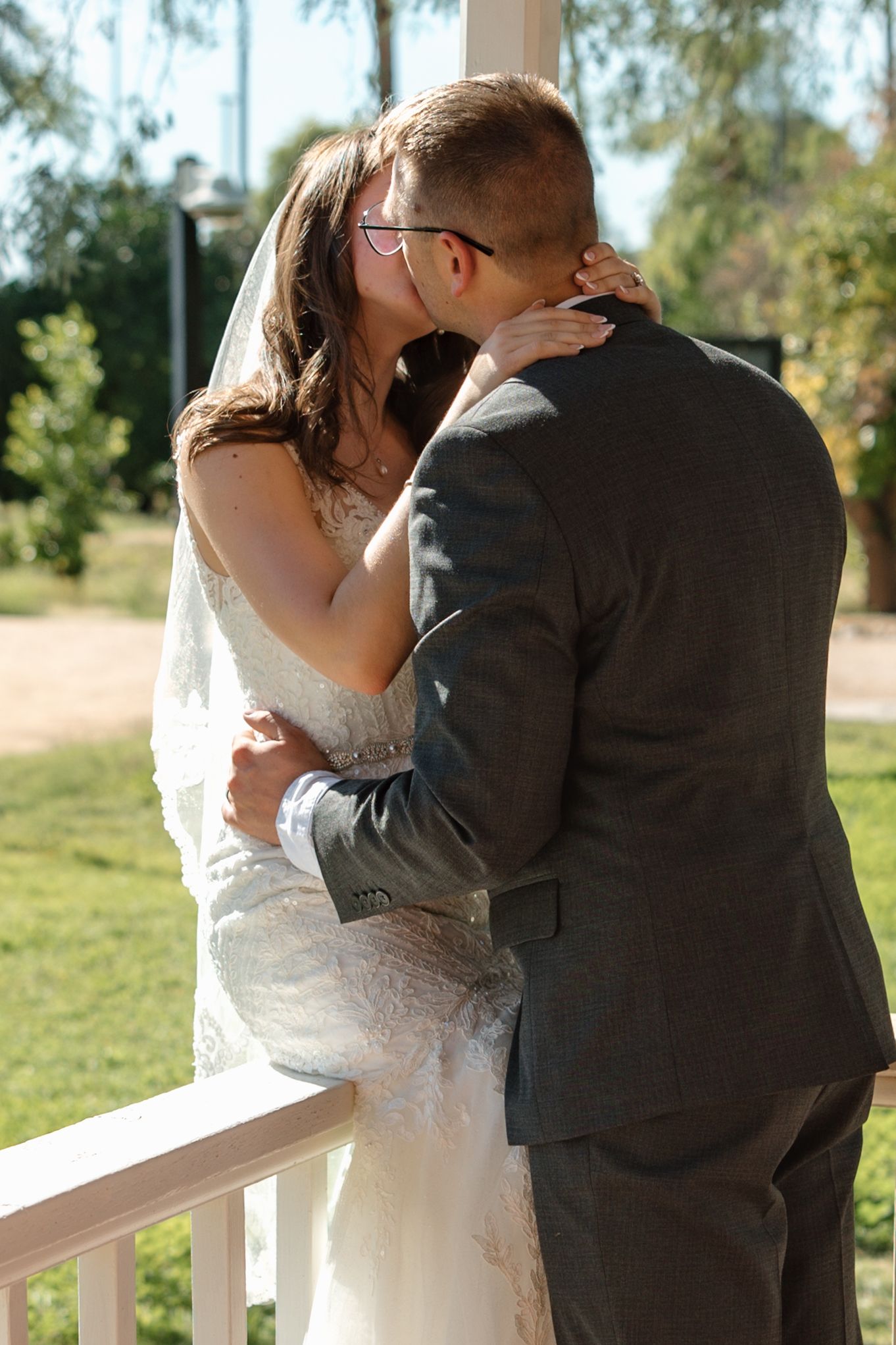 Bride and groom share a romantic first kiss on a white porch railing surrounded by lush green trees.