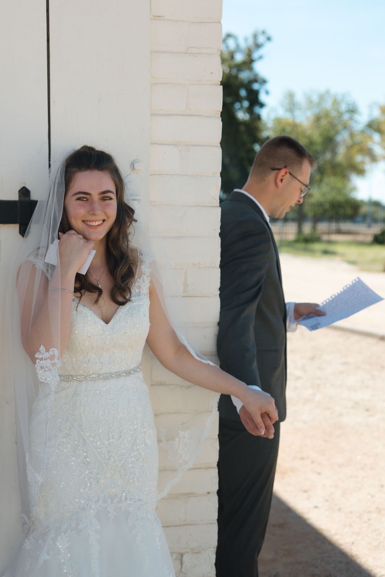 Bride in white lace gown smiles while groom in dark suit reads vows behind a white brick wall outdoors.