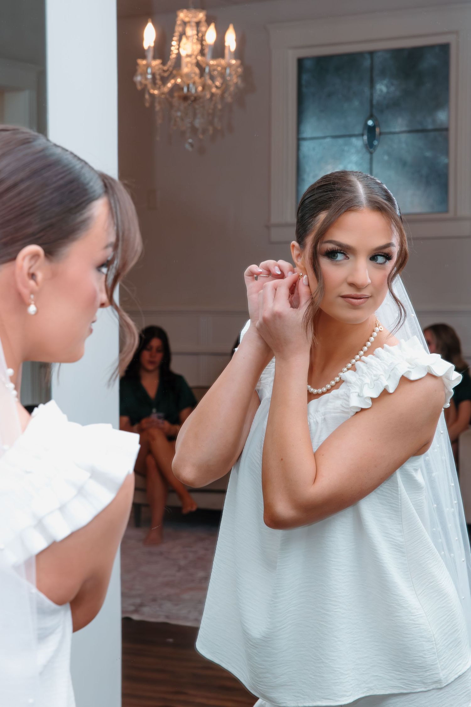 Bride in white ruffled dress adjusting earrings while looking in mirror during wedding day preparations.