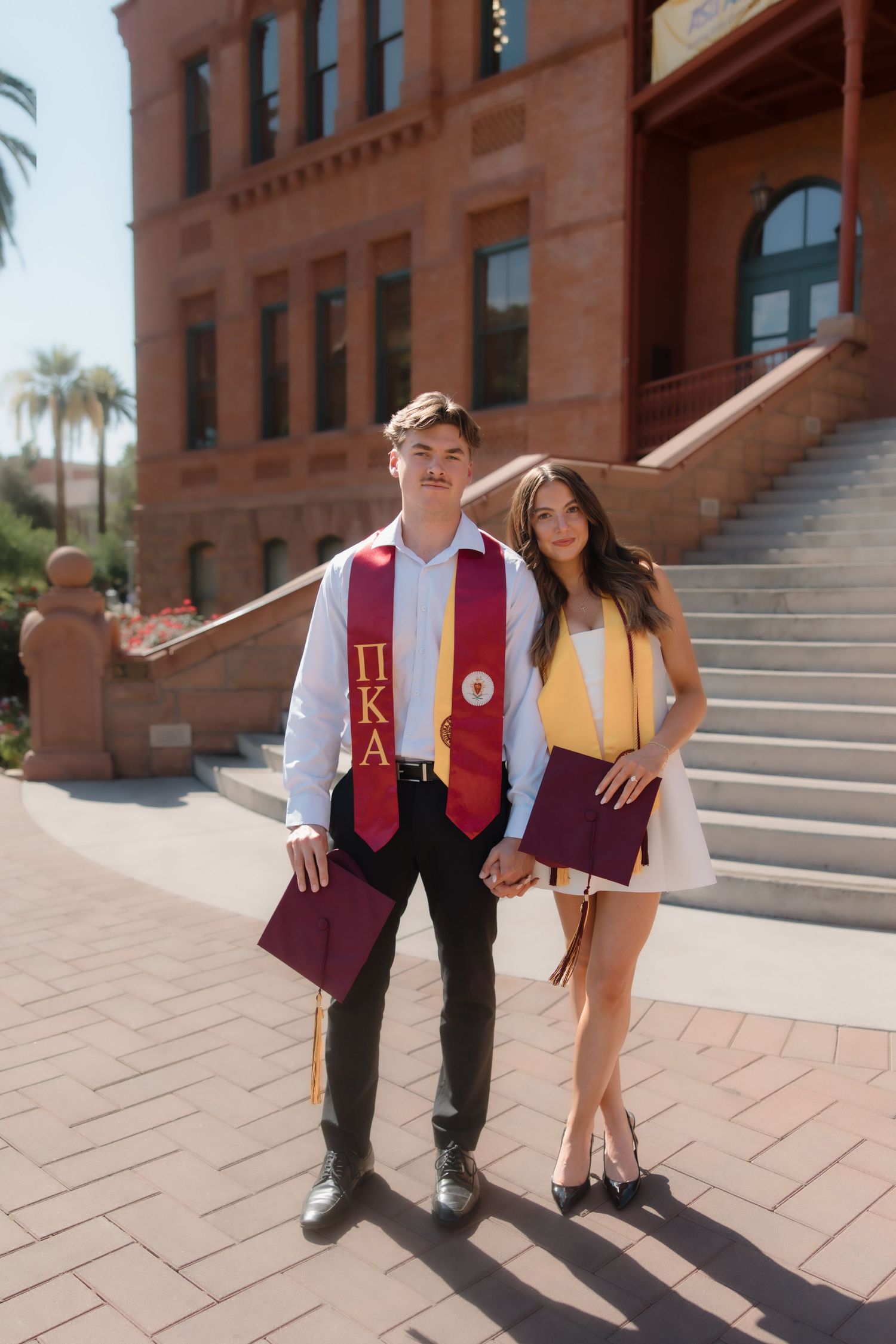 Smiling couple in graduation stoles posing together outside red brick university building on sunny day.