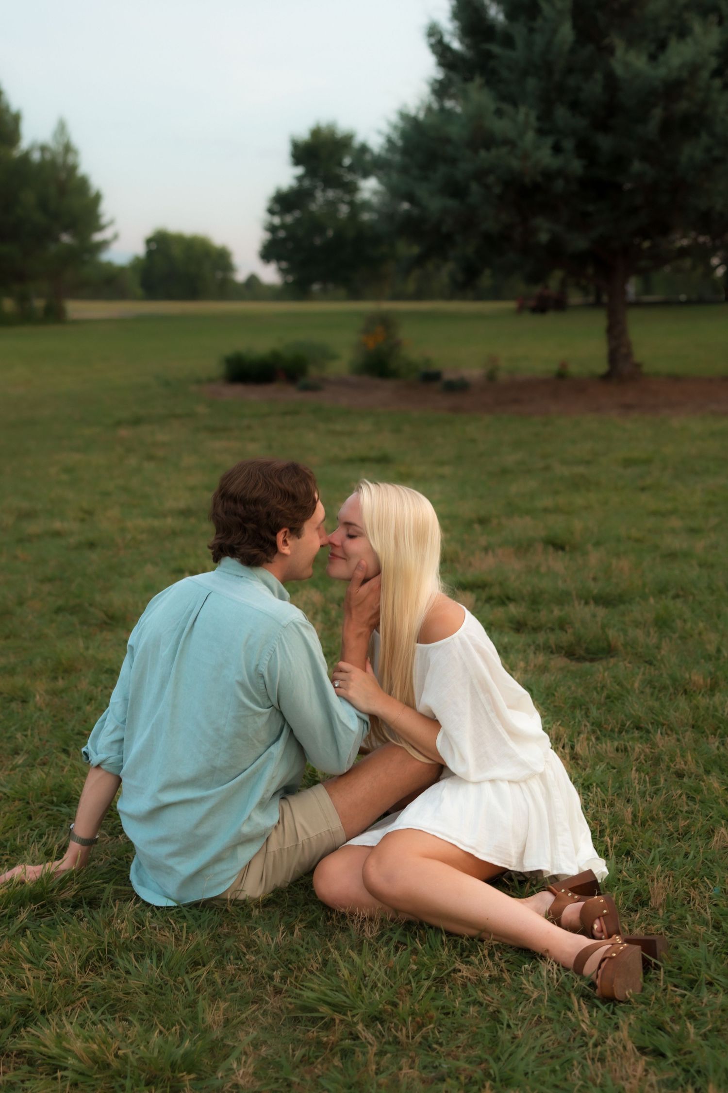 Young couple leaning in for a kiss while sitting on grass in a scenic outdoor park.