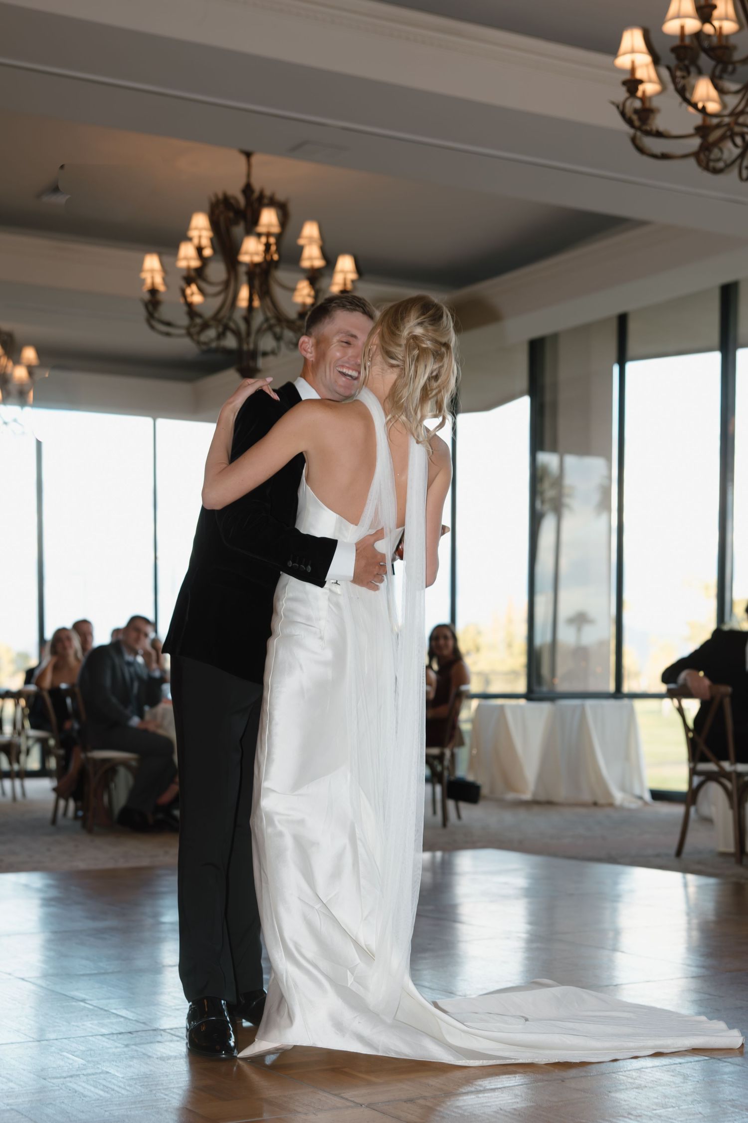 Bride and groom share a joyful first dance embrace at elegant wedding reception with chandelier lighting.