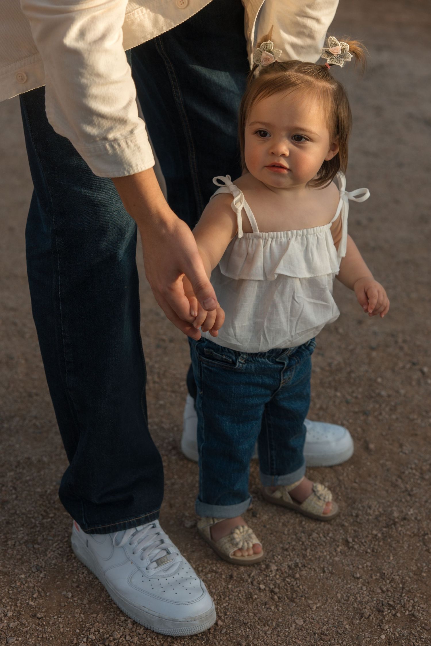 Toddler girl in white ruffled top and jeans holds parent's hand while learning to walk outdoors.