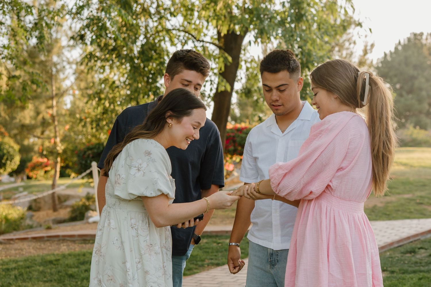 Four young adults holding hands in a circle outdoors in a sunny park setting.