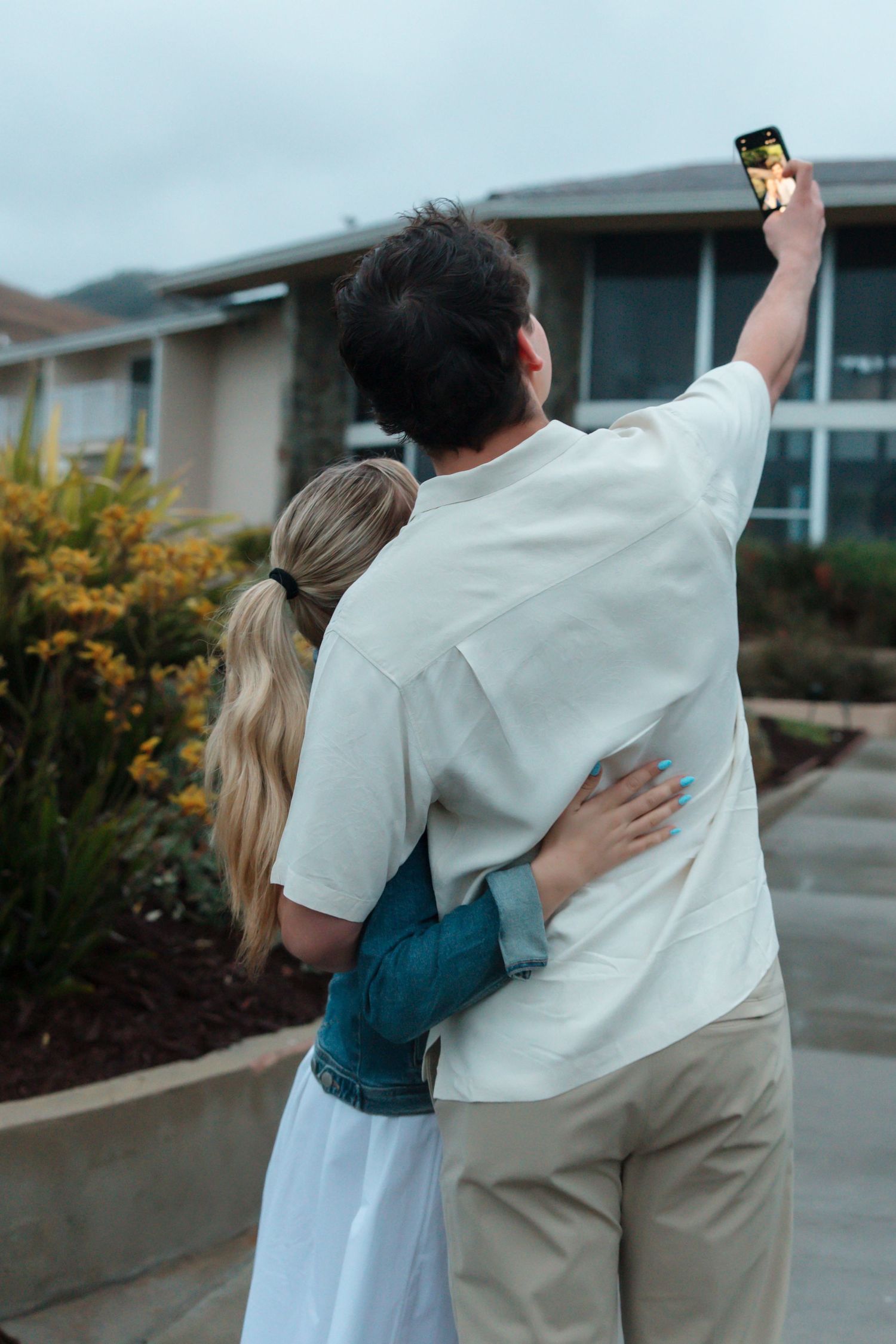 Couple taking a selfie while embracing outside a modern home surrounded by lush landscaping.