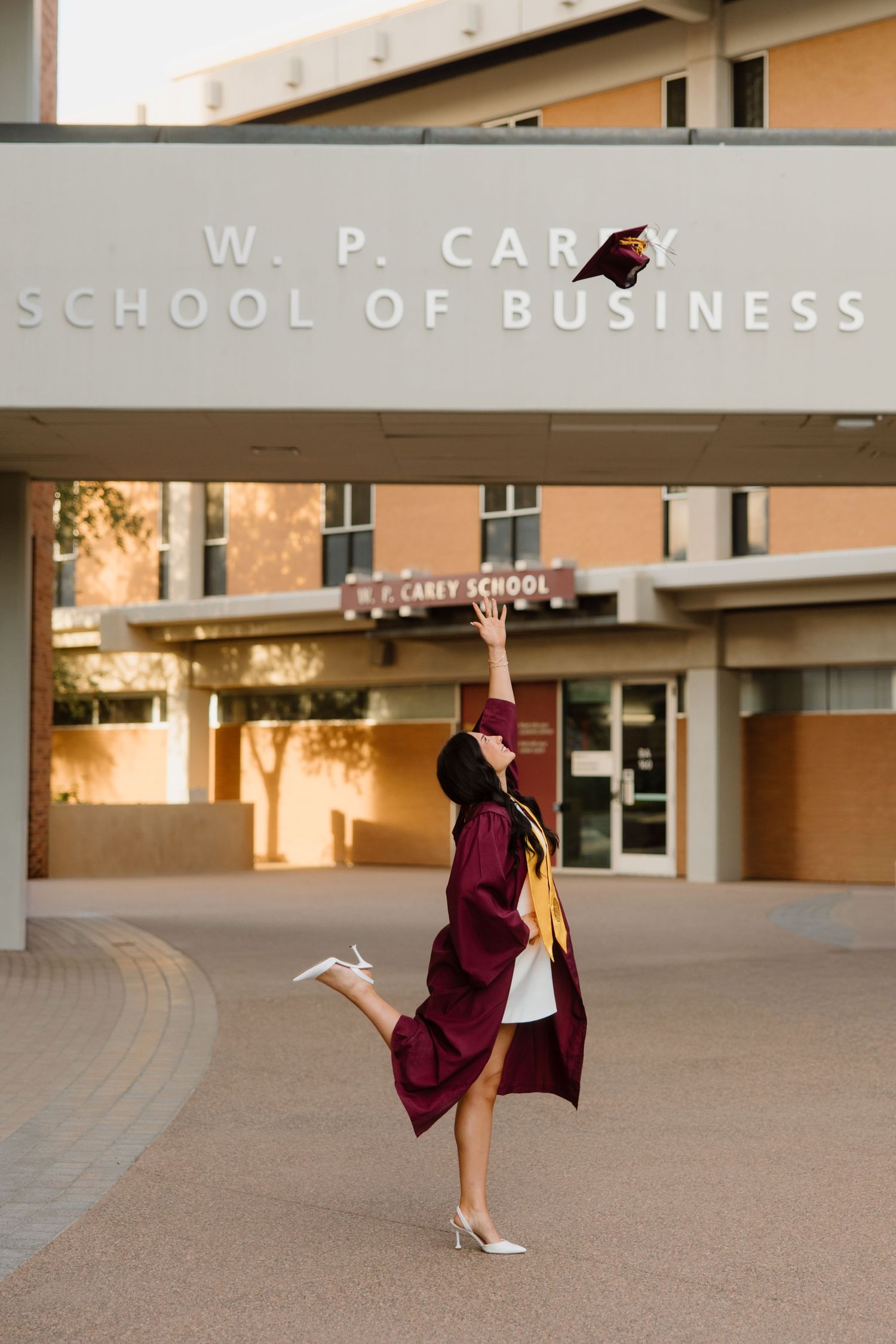 Graduate in maroon gown tosses cap in front of W.P. Carey School of Business building at Arizona State University.