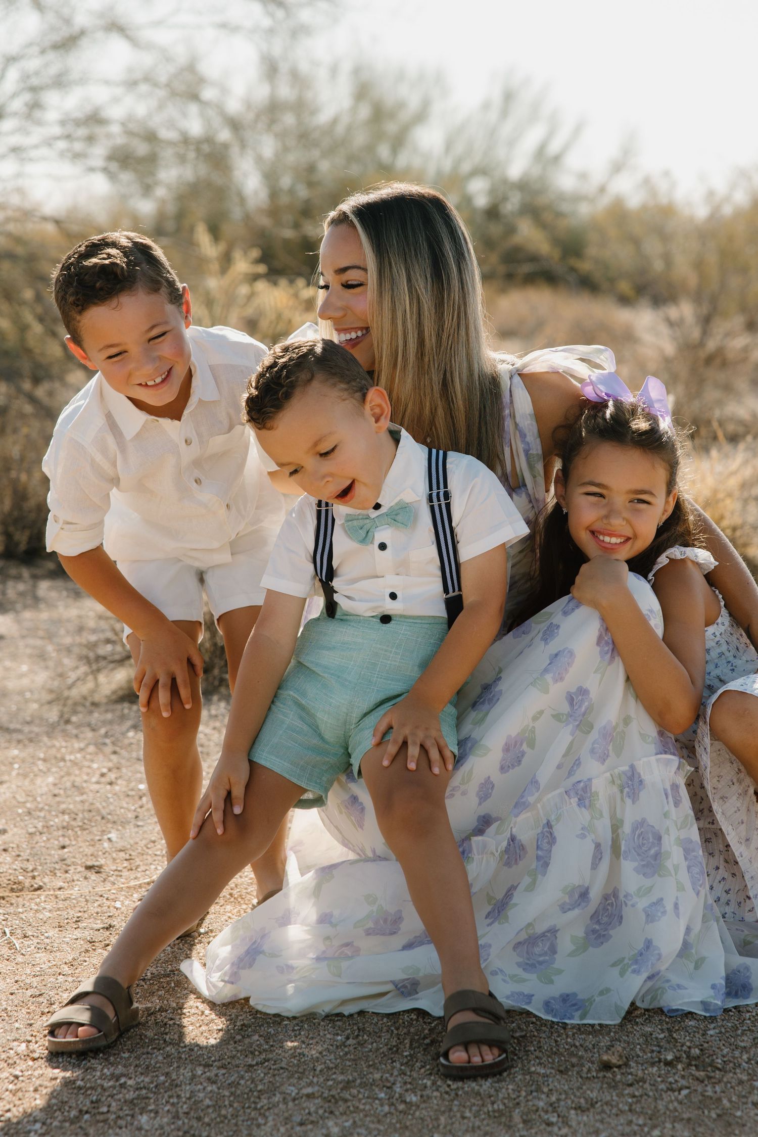 A smiling mother sits outdoors with her three young children dressed in pastel outfits, sharing a joyful family moment.