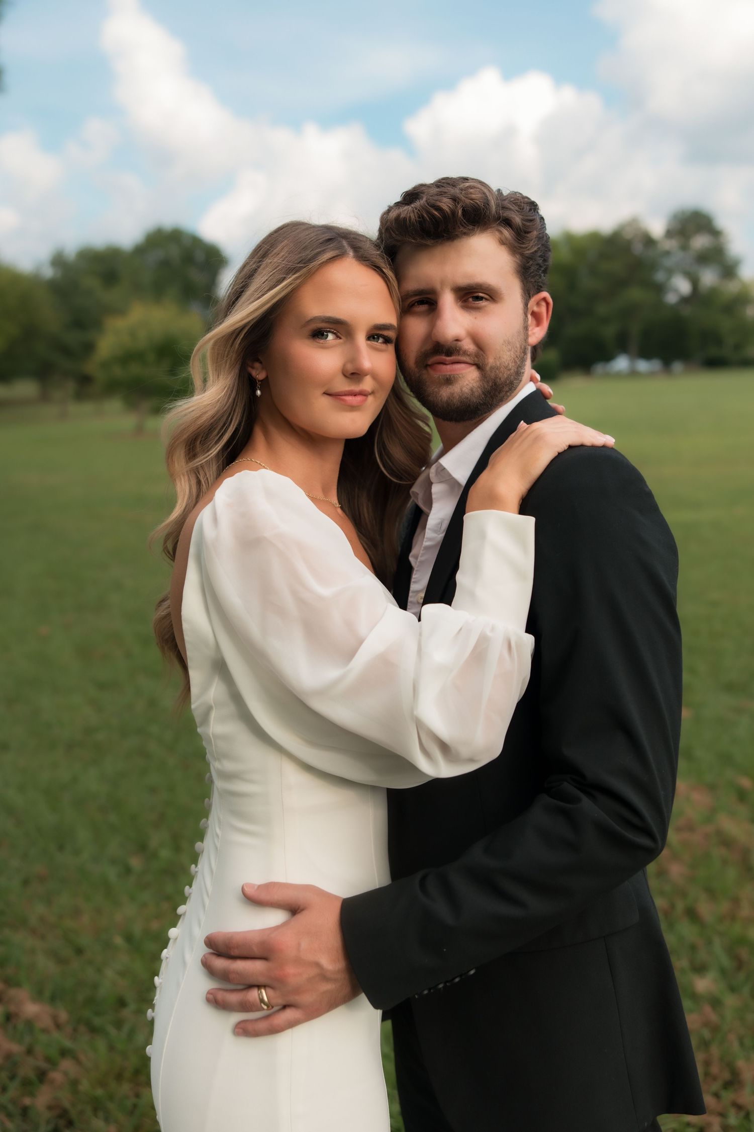 Newlywed couple embracing outdoors, bride in white long-sleeve gown and groom in black suit, smiling at camera.