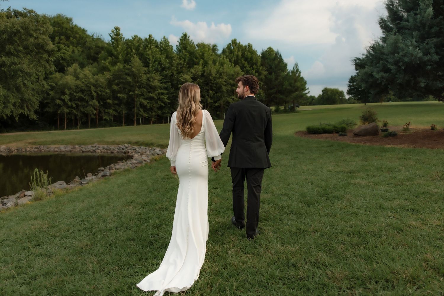 Bride and groom walk hand in hand across green lawn toward a scenic pond and trees.