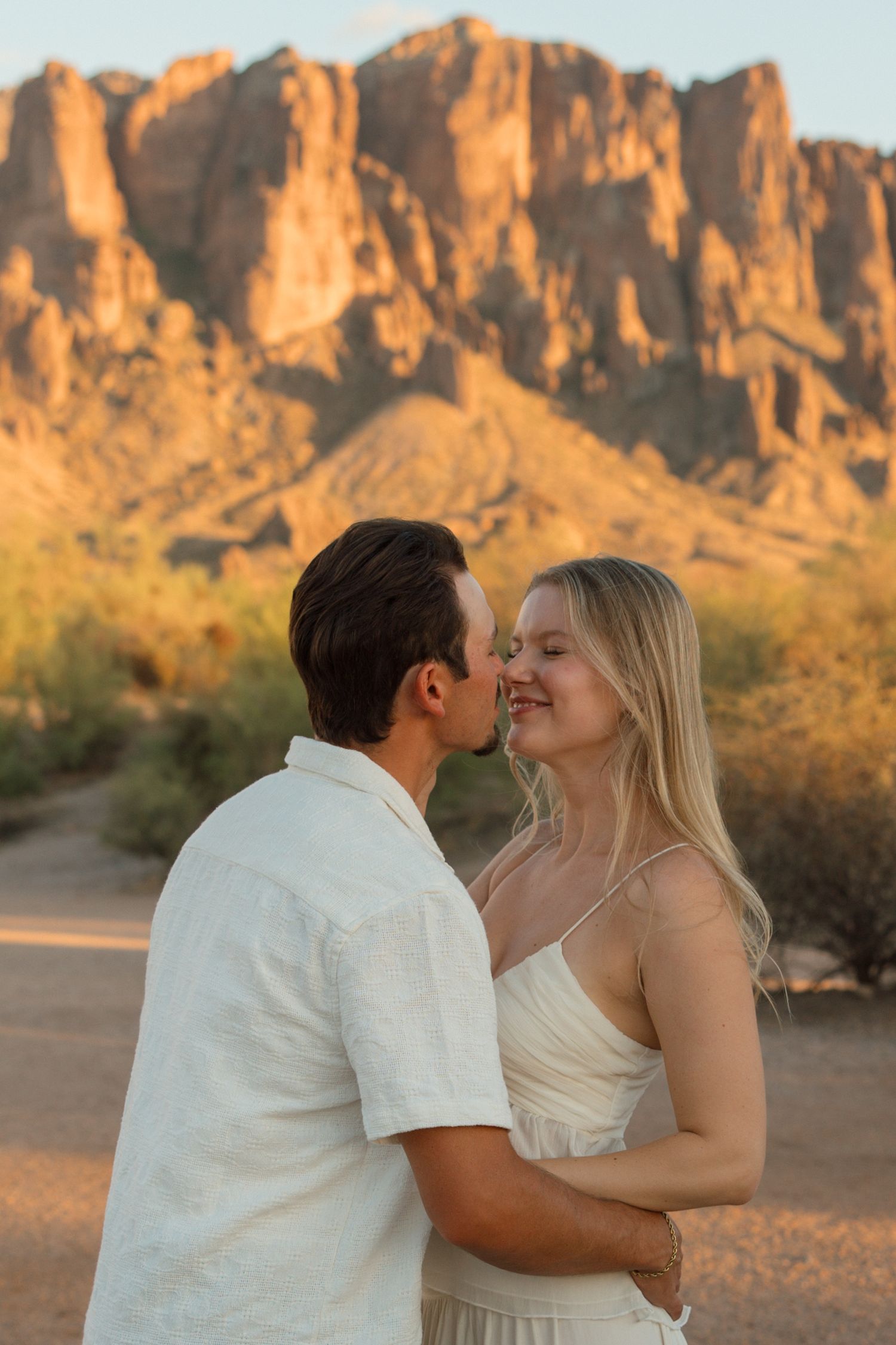 Couple sharing a romantic kiss at golden hour with Superstition Mountains in Arizona desert background.