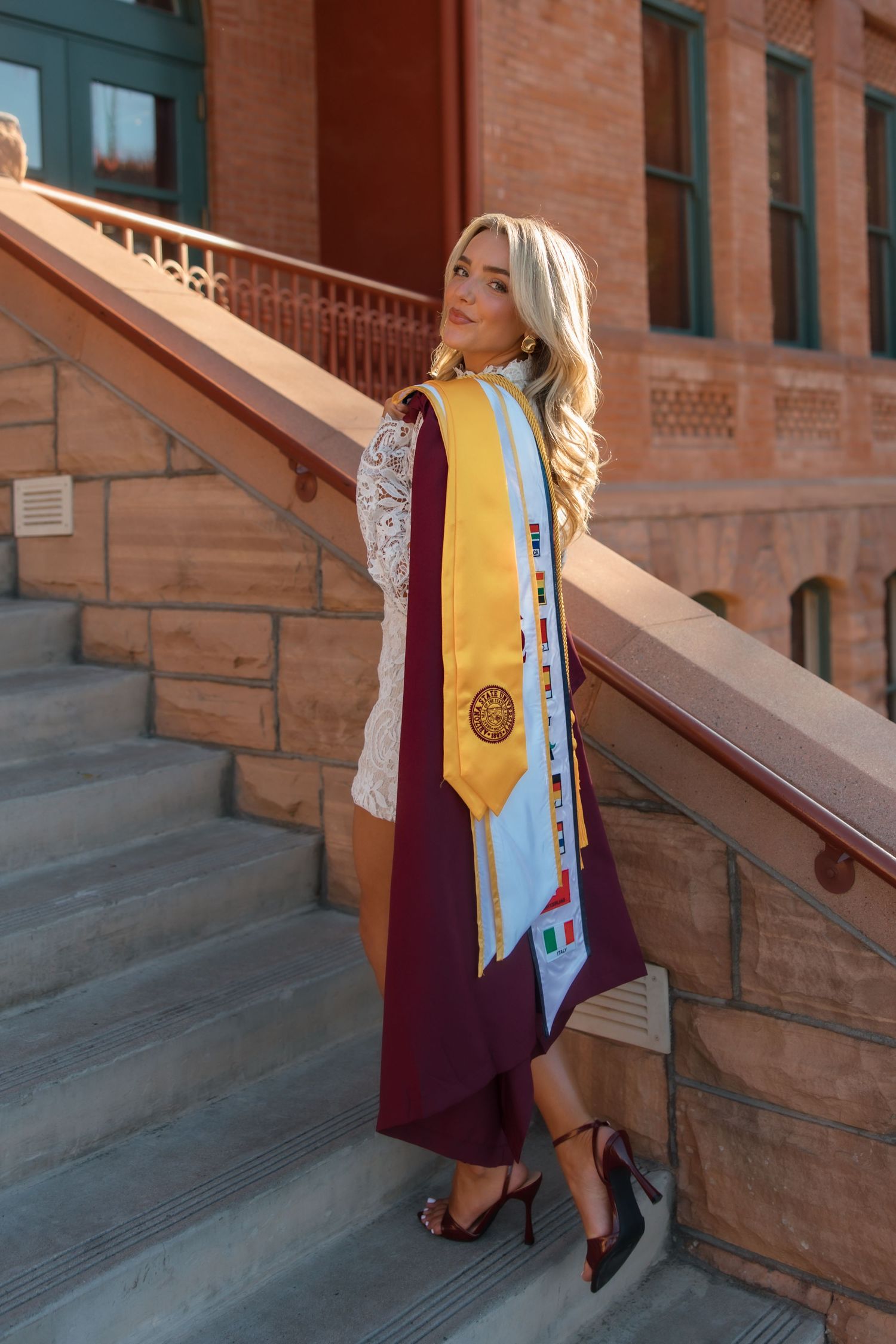 Blonde woman in graduation attire with maroon gown and gold sash posing on brick university steps.