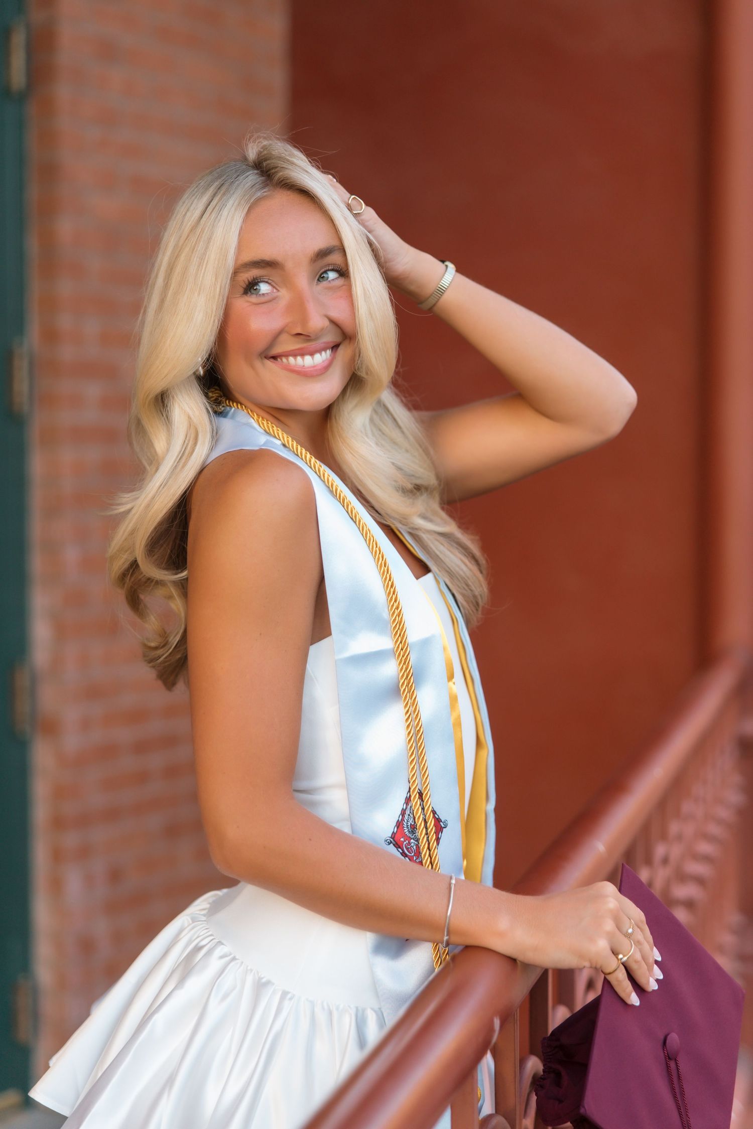 Smiling blonde graduate in white cap gown with gold honor cords posing outdoors near brick building.