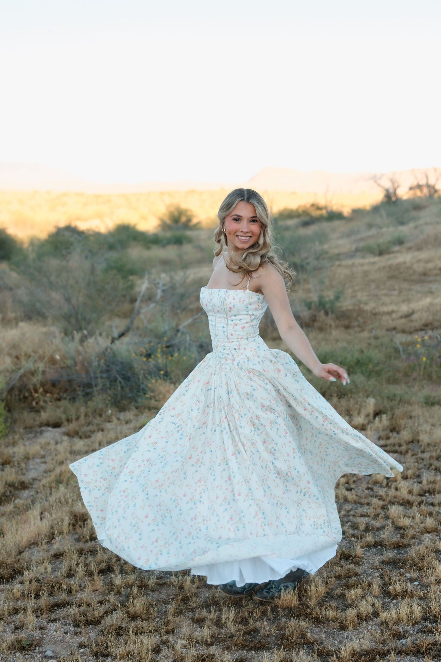 Smiling blonde woman spinning in a flowing white floral strapless dress in a rustic desert field at dusk.