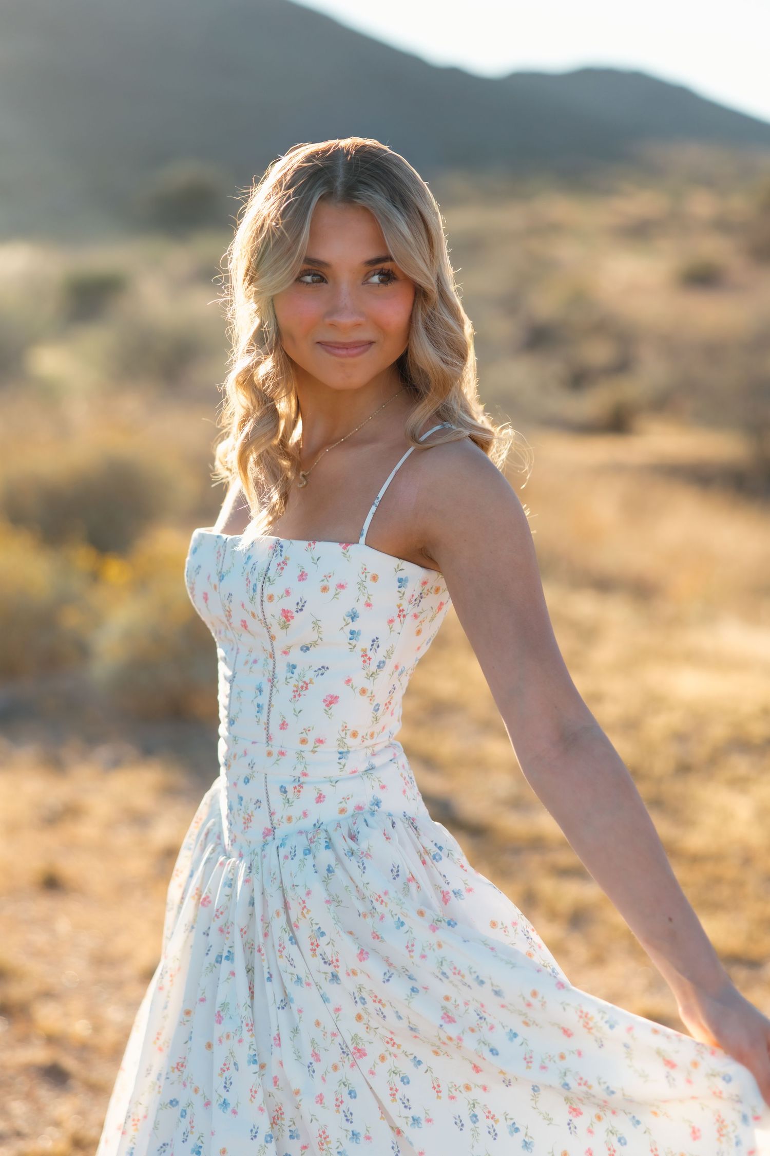 Young woman in white floral sundress poses gracefully in a sunny desert landscape with golden hills in the background.