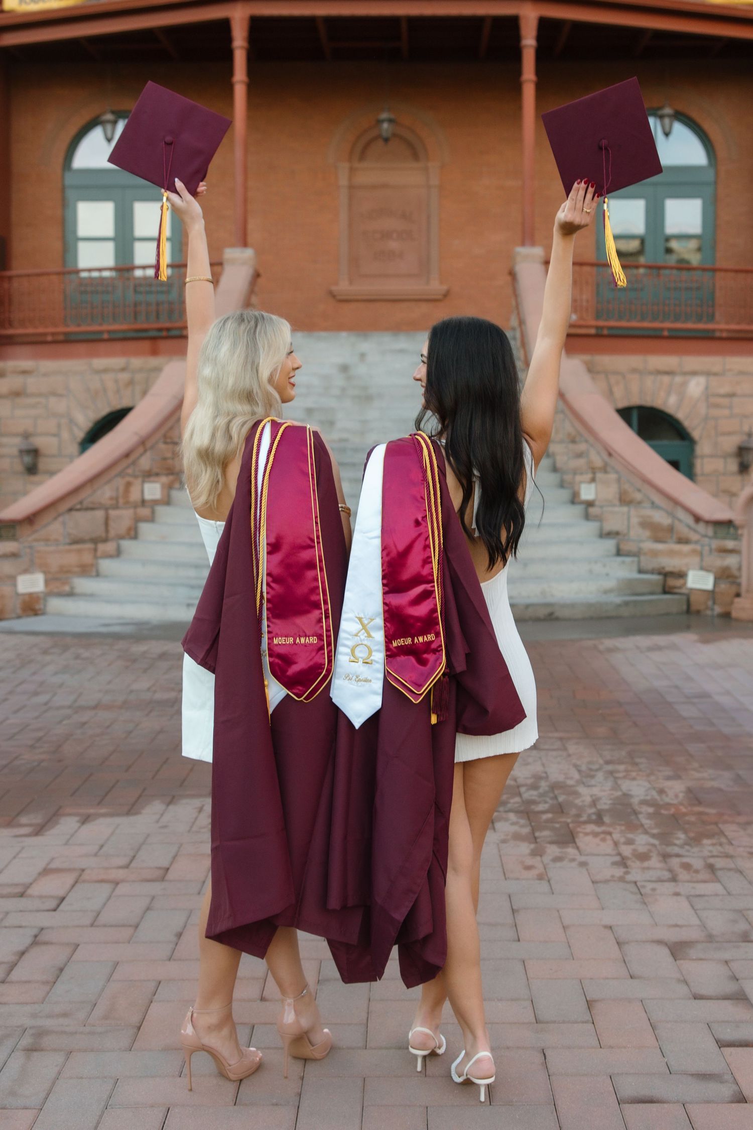 Two ASU graduates in maroon robes tossing caps in the air, celebrating commencement outside a historic building.