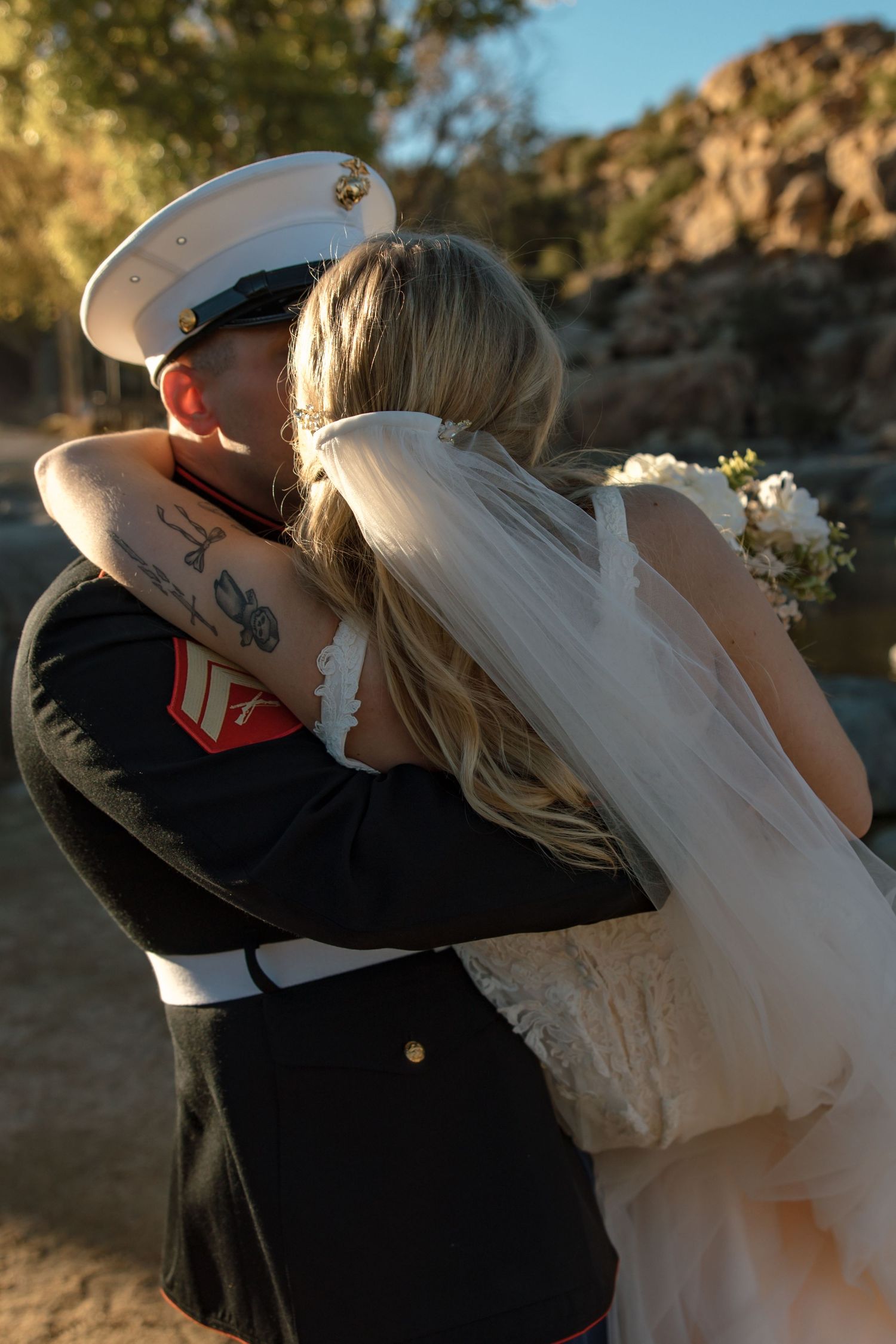Marine groom embraces bride in elegant gown with veil in warm golden outdoor lighting.