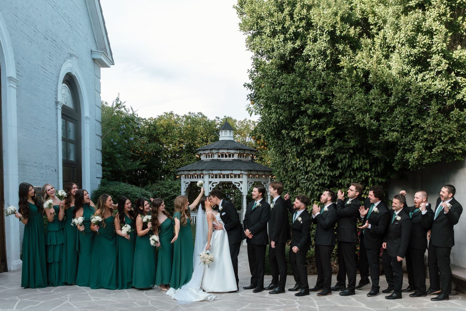 Large wedding party poses with bride and groom kissing in front of a white gazebo outdoors.