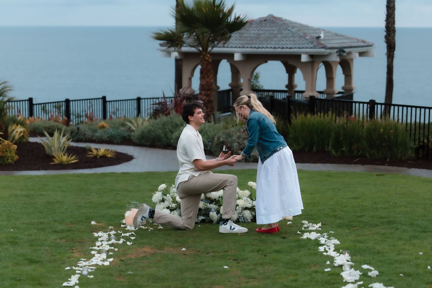 Man kneeling on grass proposing to woman with ocean gazebo backdrop and white flower arrangement.