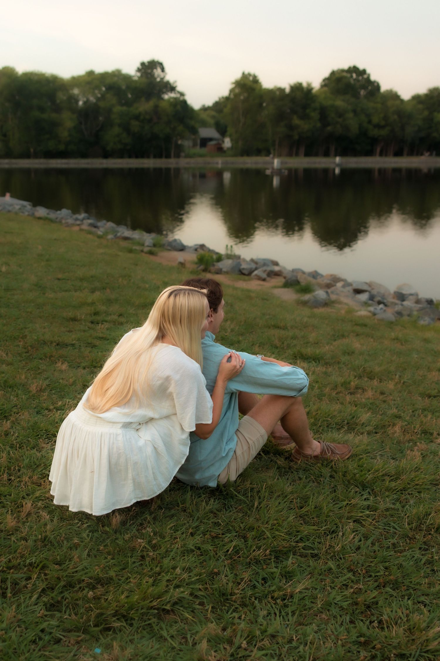 Young couple sitting closely together on grass overlooking a calm lake at dusk.