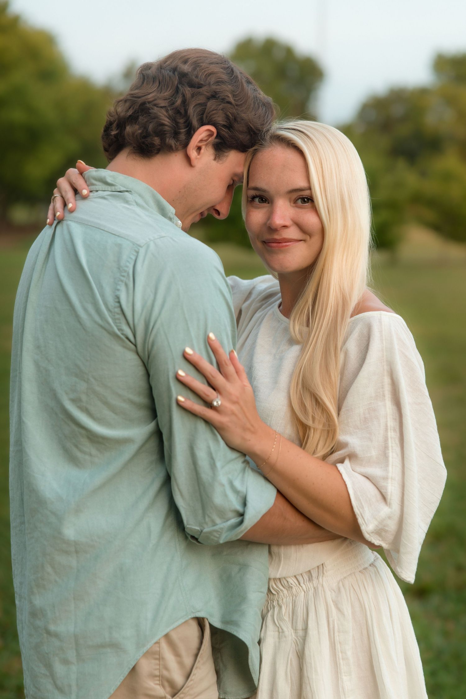 Blonde woman smiles at camera while embracing her partner in a green park.