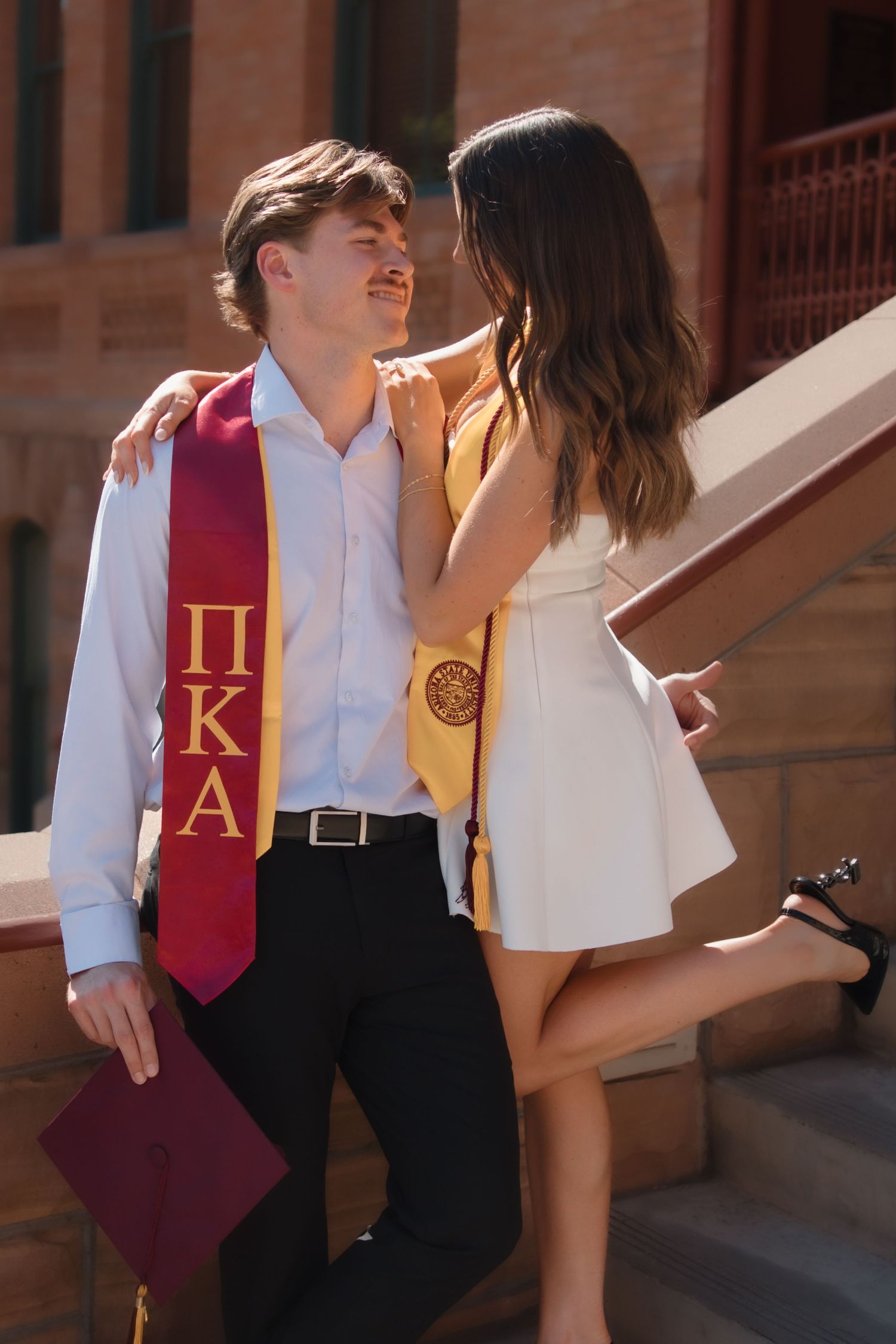 College graduates in Pi Kappa Alpha and gold honor stoles sharing romantic moment on brick campus steps.