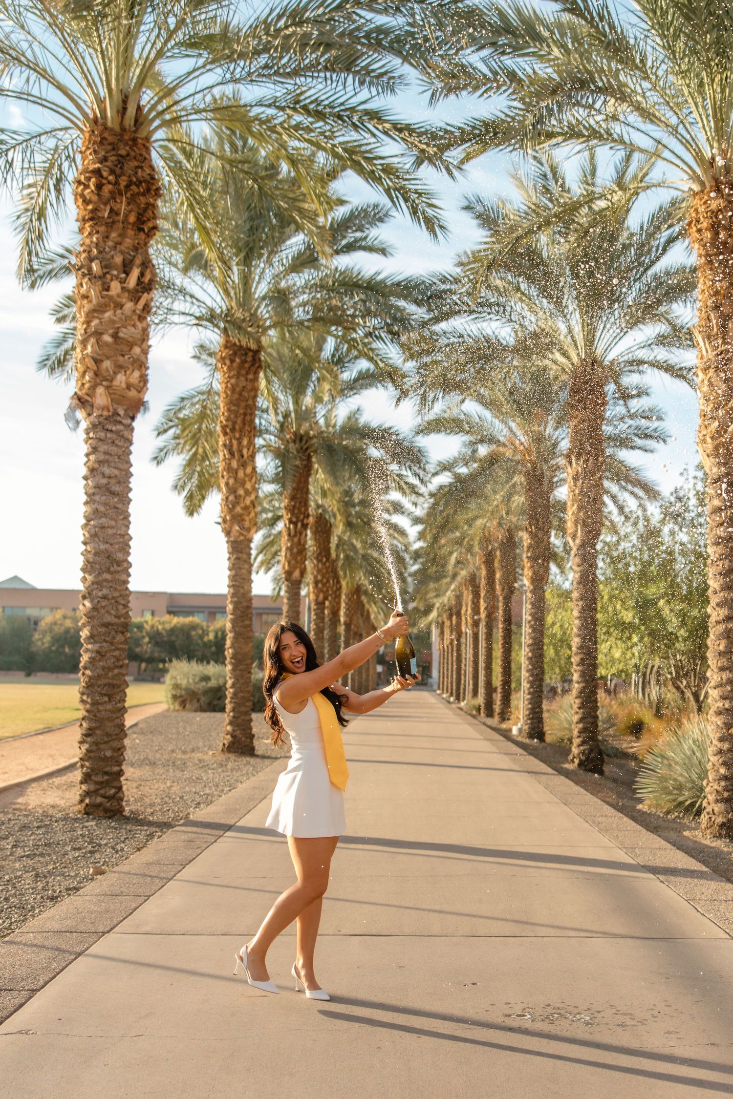 A young woman in a white dress poses joyfully on a palm tree-lined walkway in a sunny resort setting.