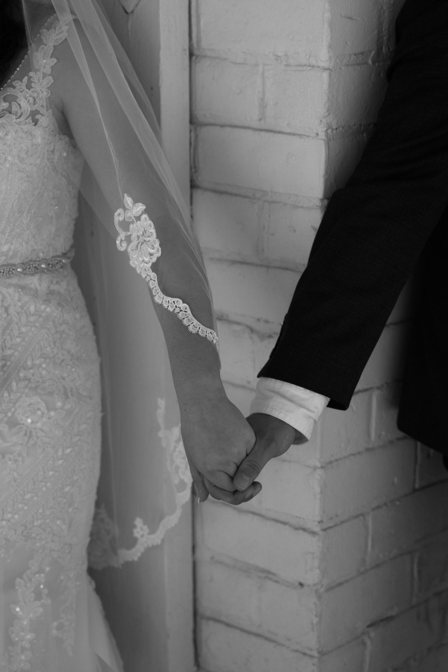 Black and white photo of bride and groom holding hands, bride wearing lace wedding dress with embroidered veil.