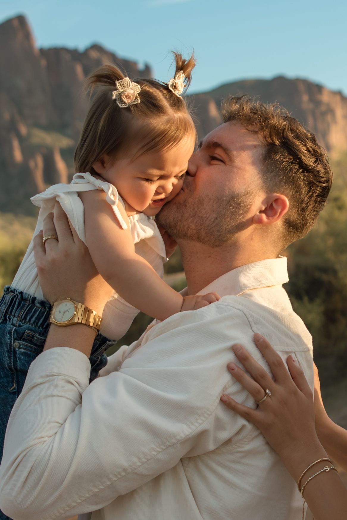 Father tenderly kissing his laughing toddler daughter against a scenic desert mountain backdrop.
