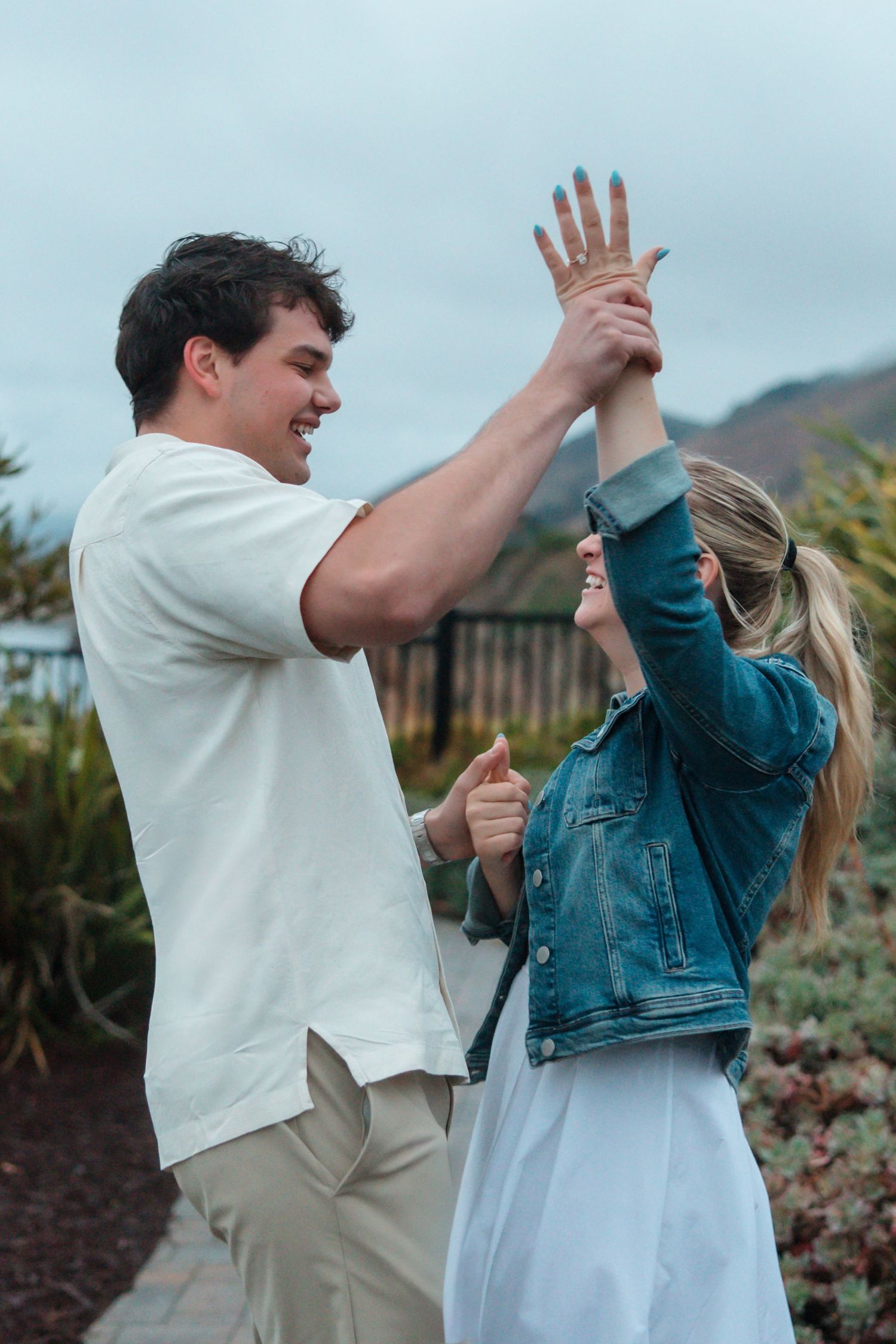 Couple laughing and raising hands together in garden with ocean and mountains behind them.