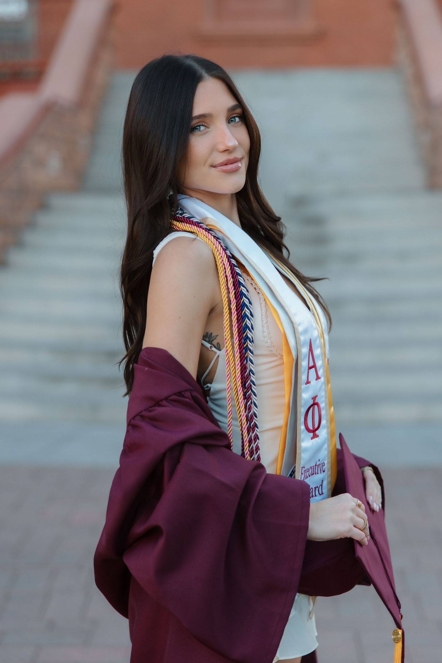Brunette woman in maroon graduation gown with Alpha Phi Executive Board stole poses on steps.