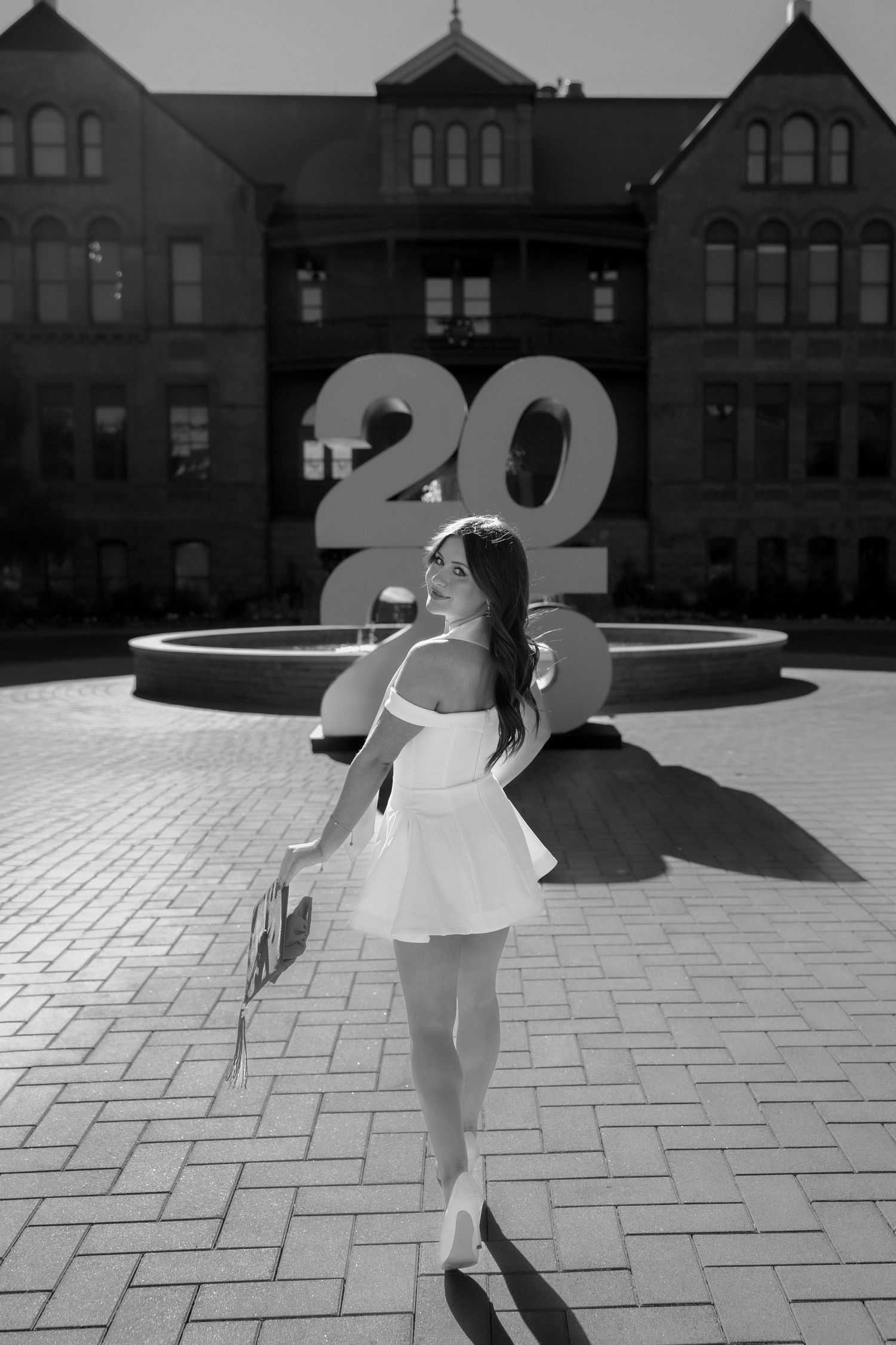 Black and white photo of a woman in white dress posing near a fountain with a '20' sign at Arizona State University.