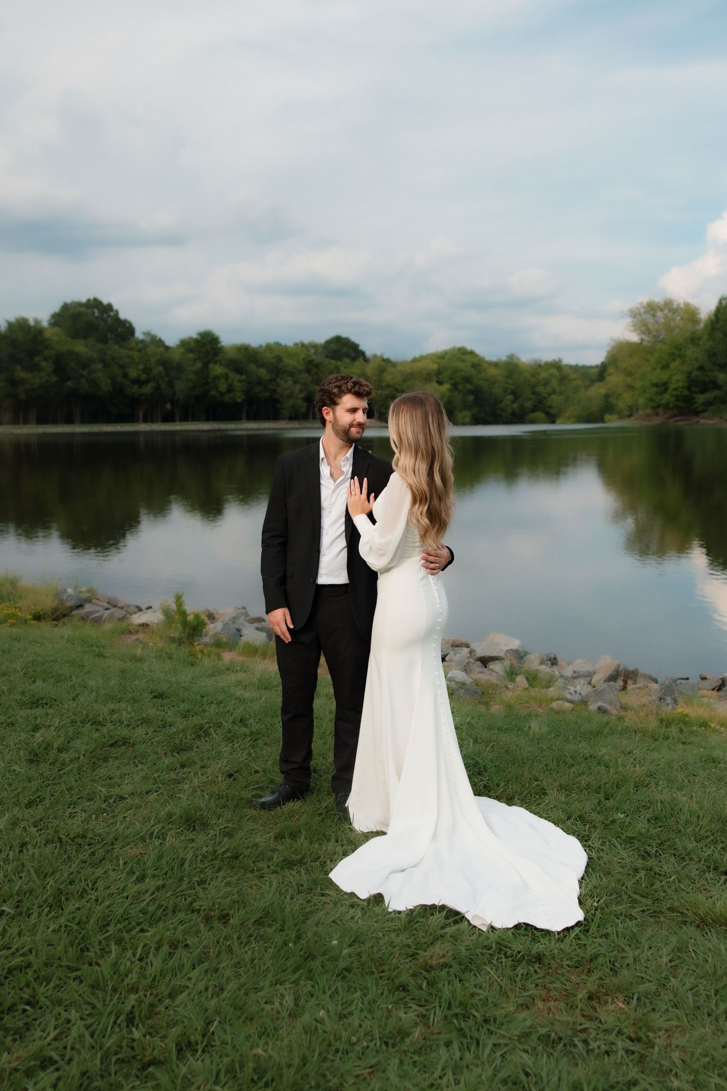 Bride and groom share a romantic moment by a peaceful lakeside, dressed in wedding attire.