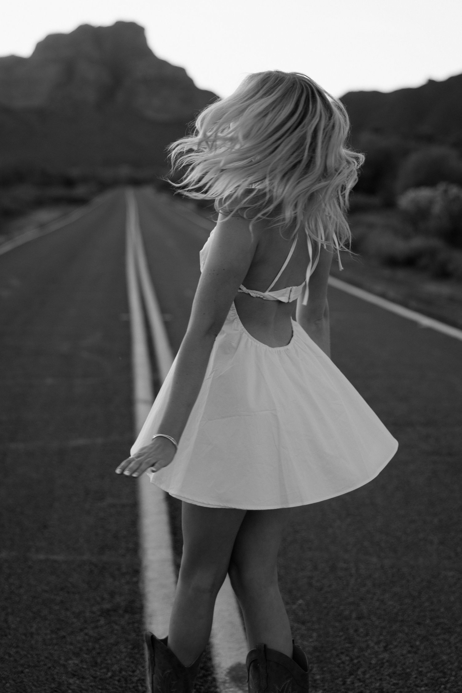Black and white photo of a blonde woman in a white mini dress and cowboy boots twirling on an open desert road.