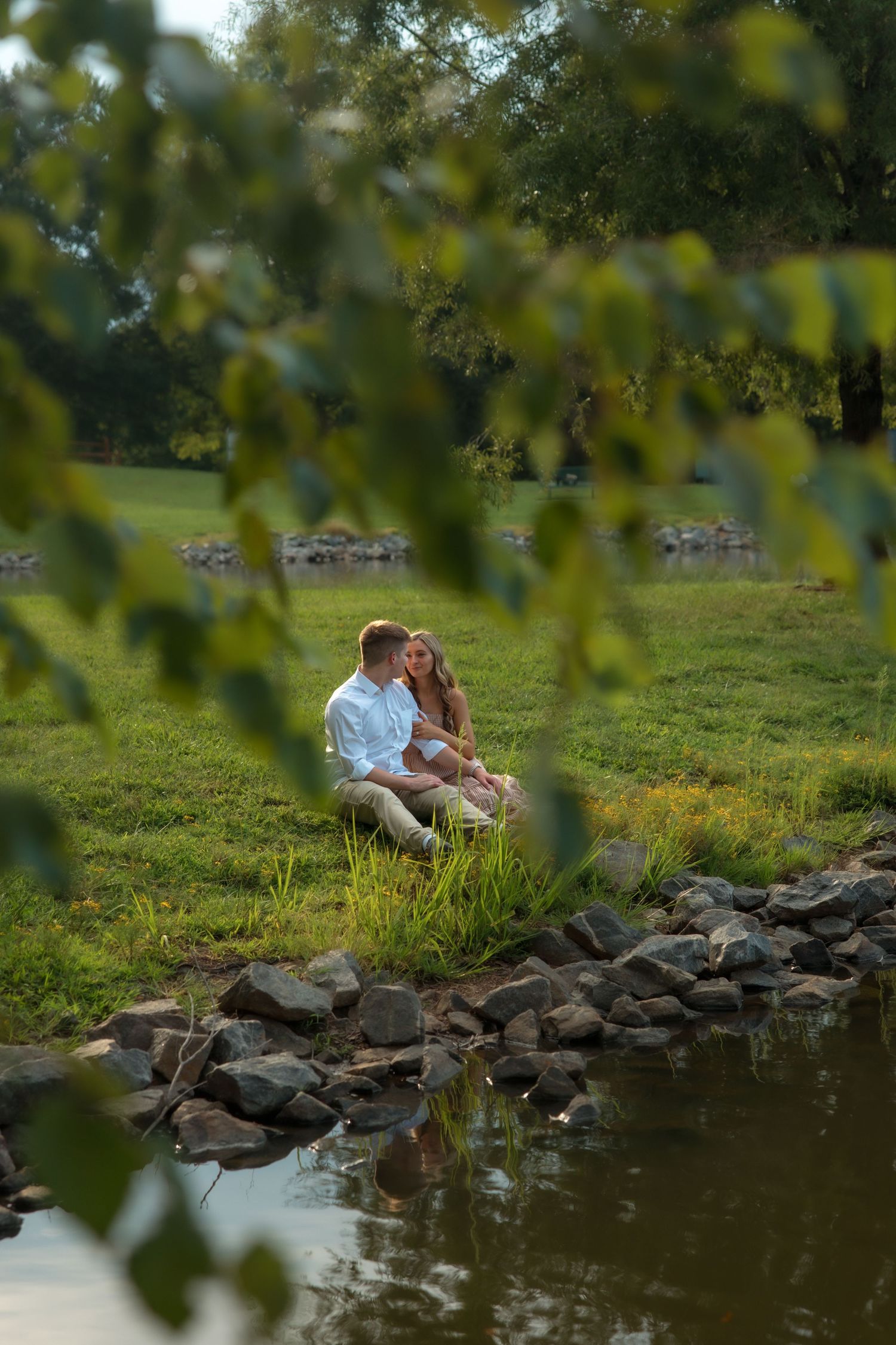Romantic couple sitting together by a rocky stream in a lush green park, framed by leafy branches.