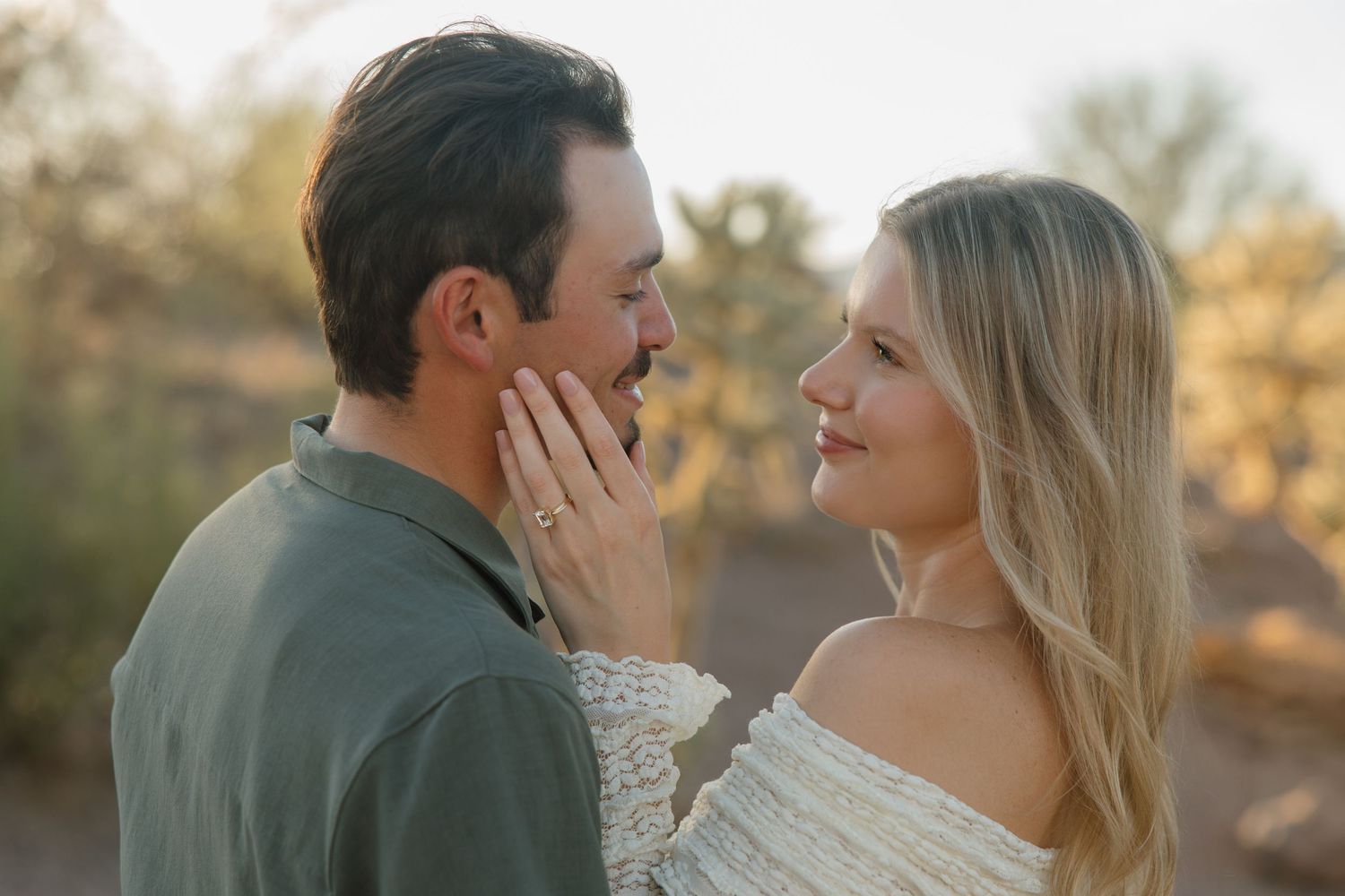Couple sharing a romantic moment outdoors at golden hour, woman wearing engagement ring touching mans face.