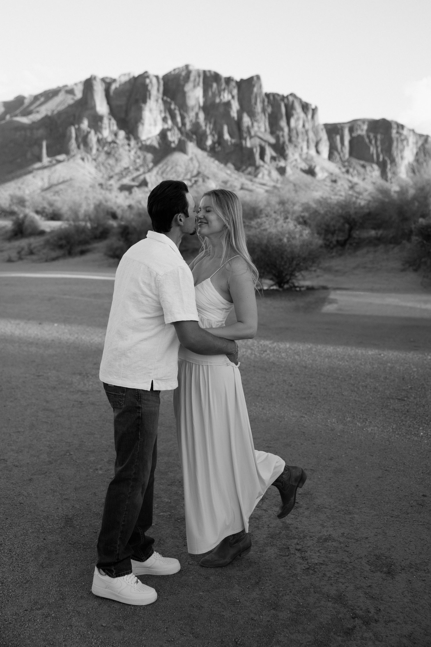 Black and white photo of a couple embracing and kissing near Superstition Mountains in Arizona desert landscape.