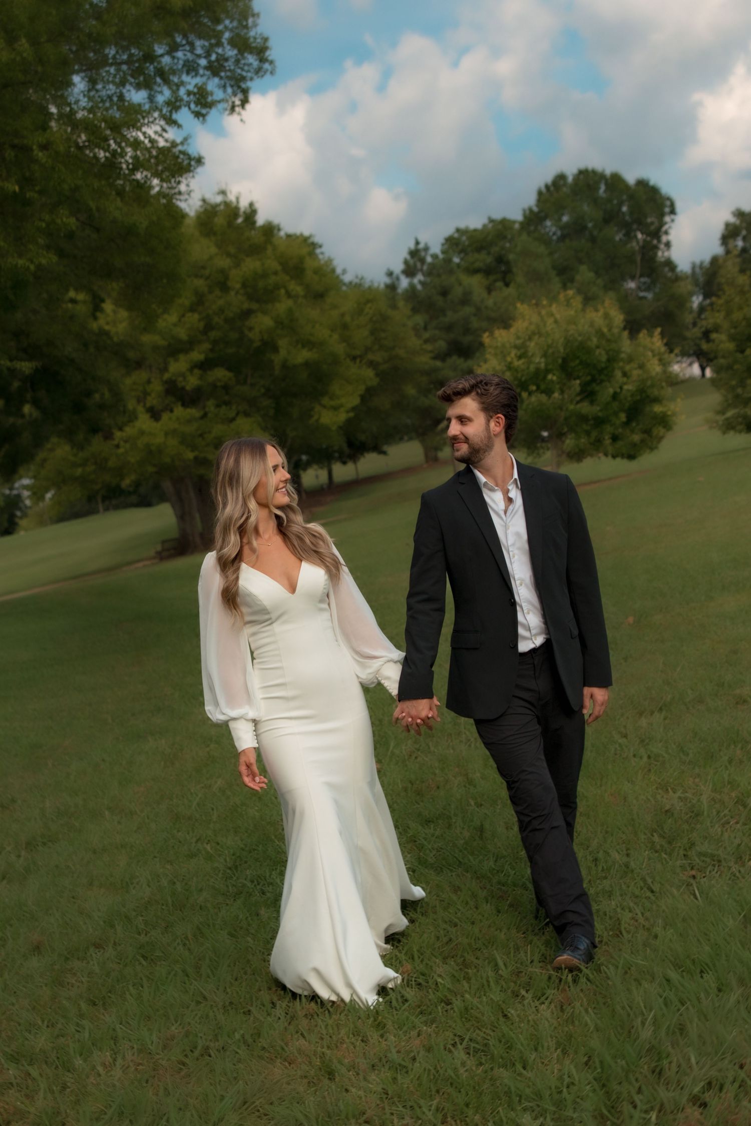 A bride in a white gown and groom in a black suit walk hand-in-hand across a lush green lawn.