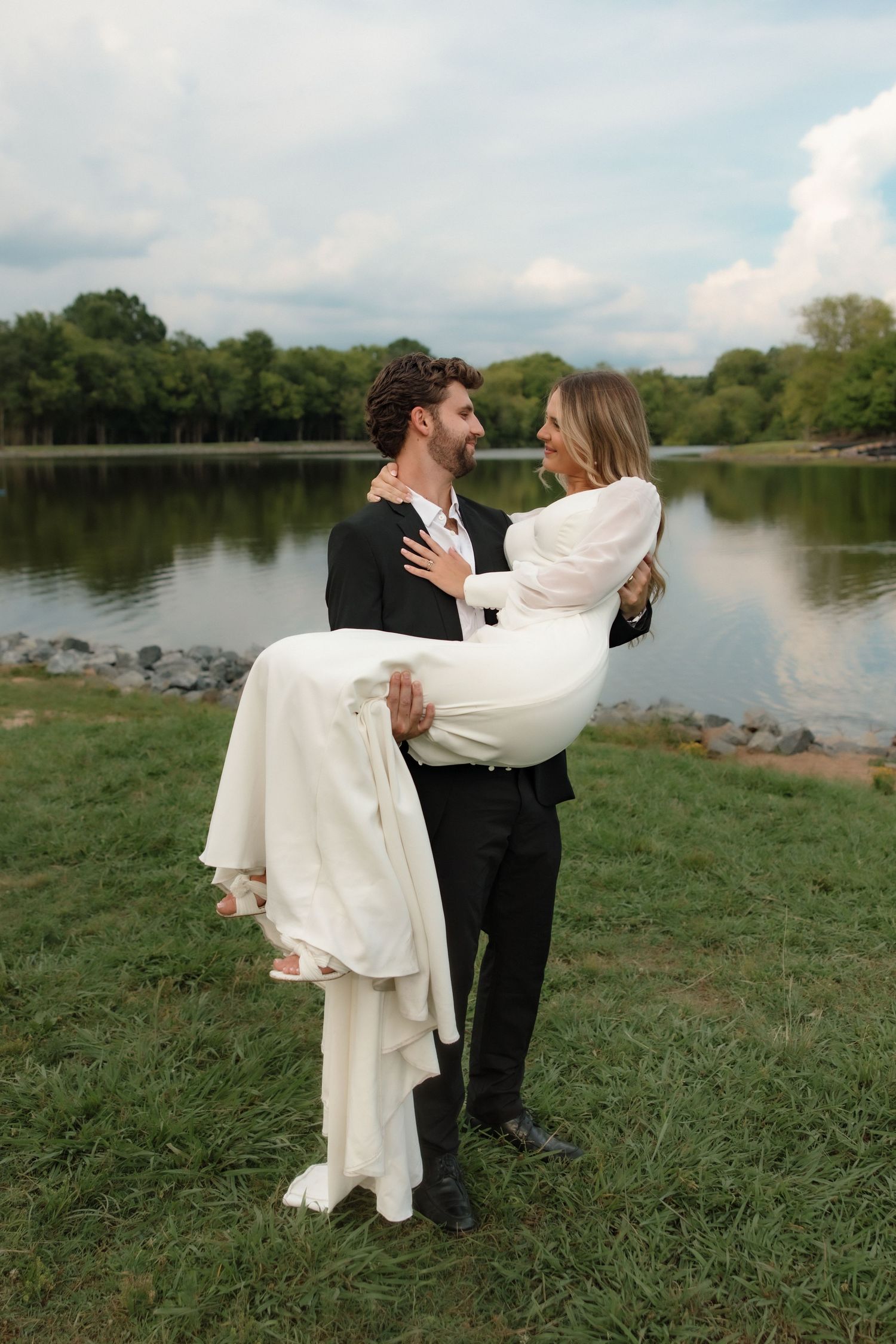 Groom carries bride in white dress by a serene lake on their wedding day outdoors.