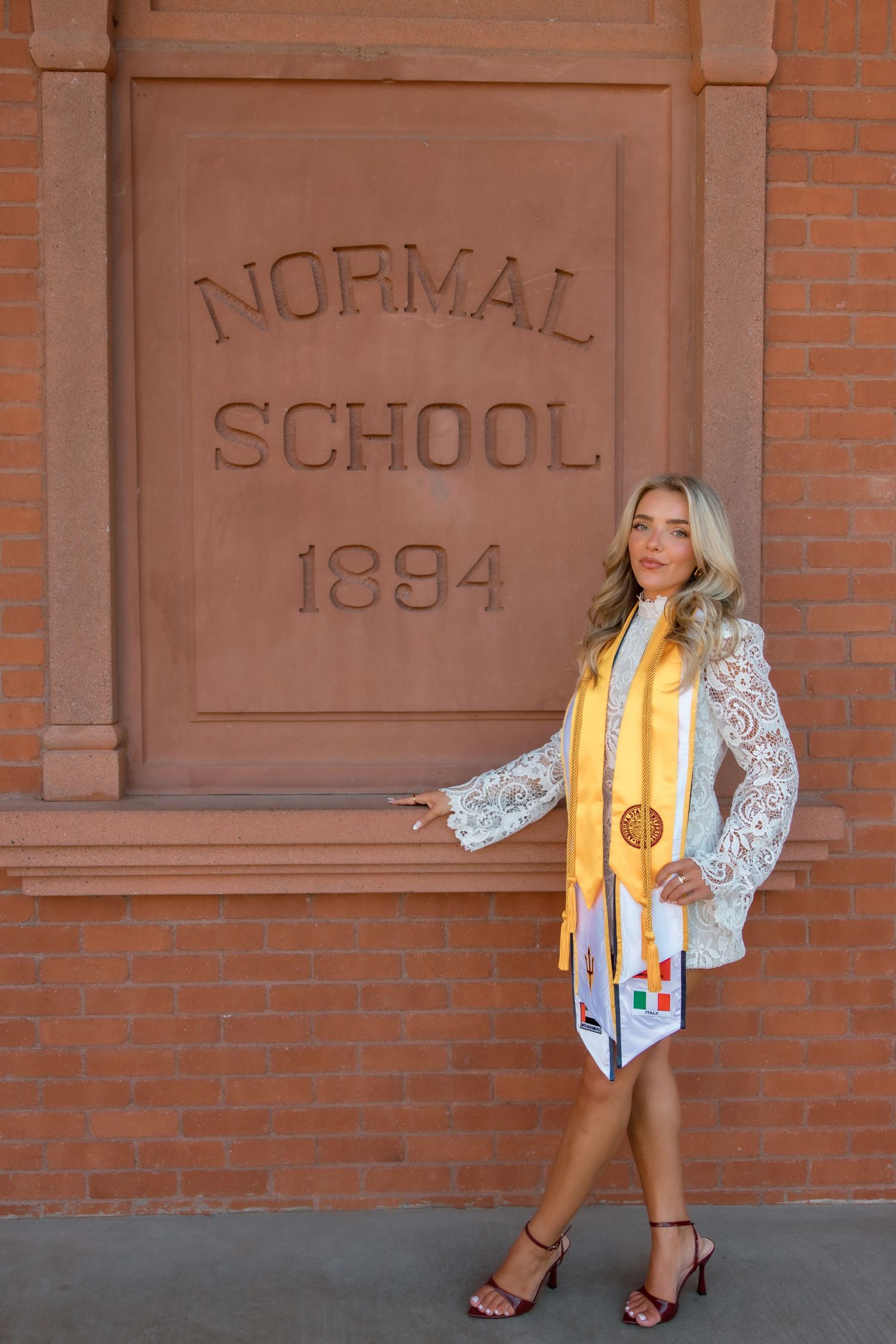 Blonde woman in white lace dress and gold graduation stole posing by Normal School 1894 brick building.