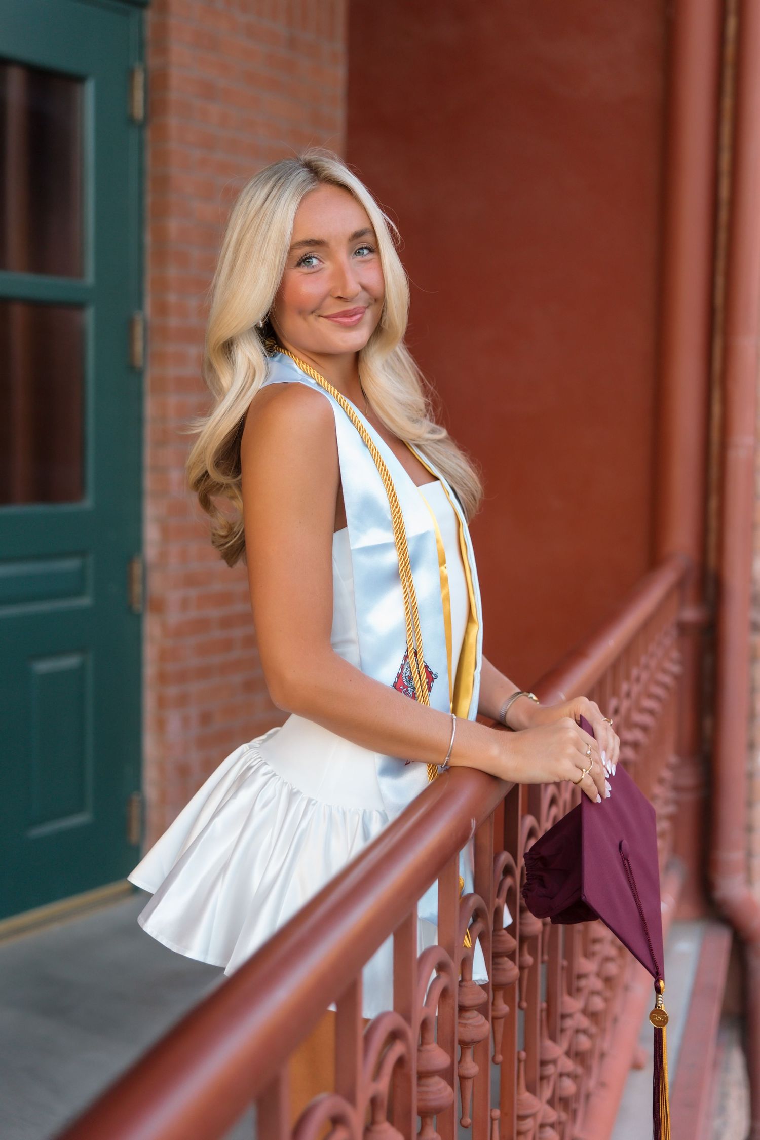 Smiling blonde graduate in white dress holding maroon cap with gold honor stole on decorative railing.