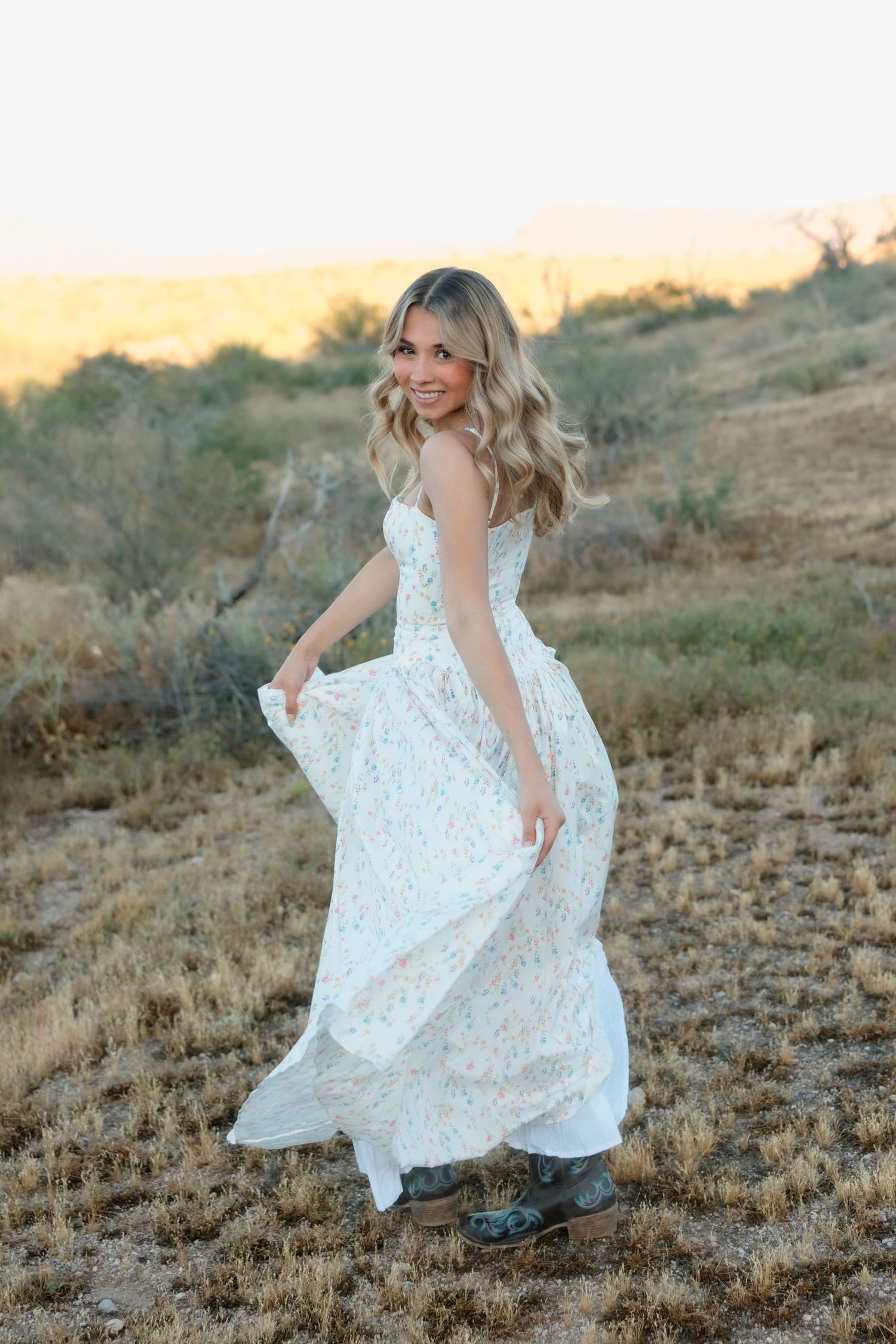 Blonde woman twirling in a white floral maxi dress with cowboy boots in a golden desert landscape at sunset.