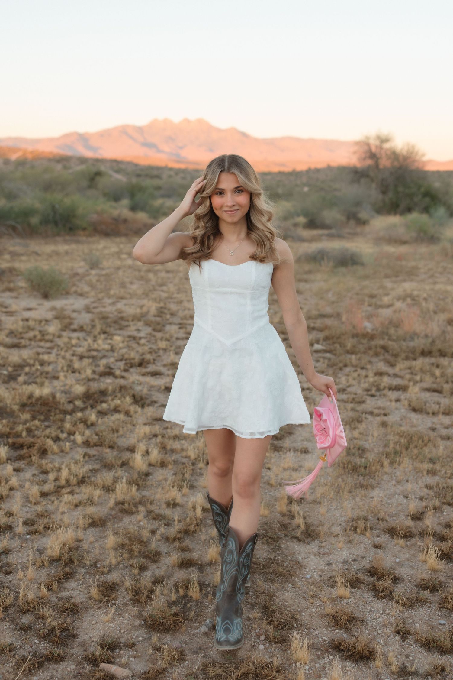 Young woman in white mini dress and cowboy boots holding pink hat in desert landscape at sunset.