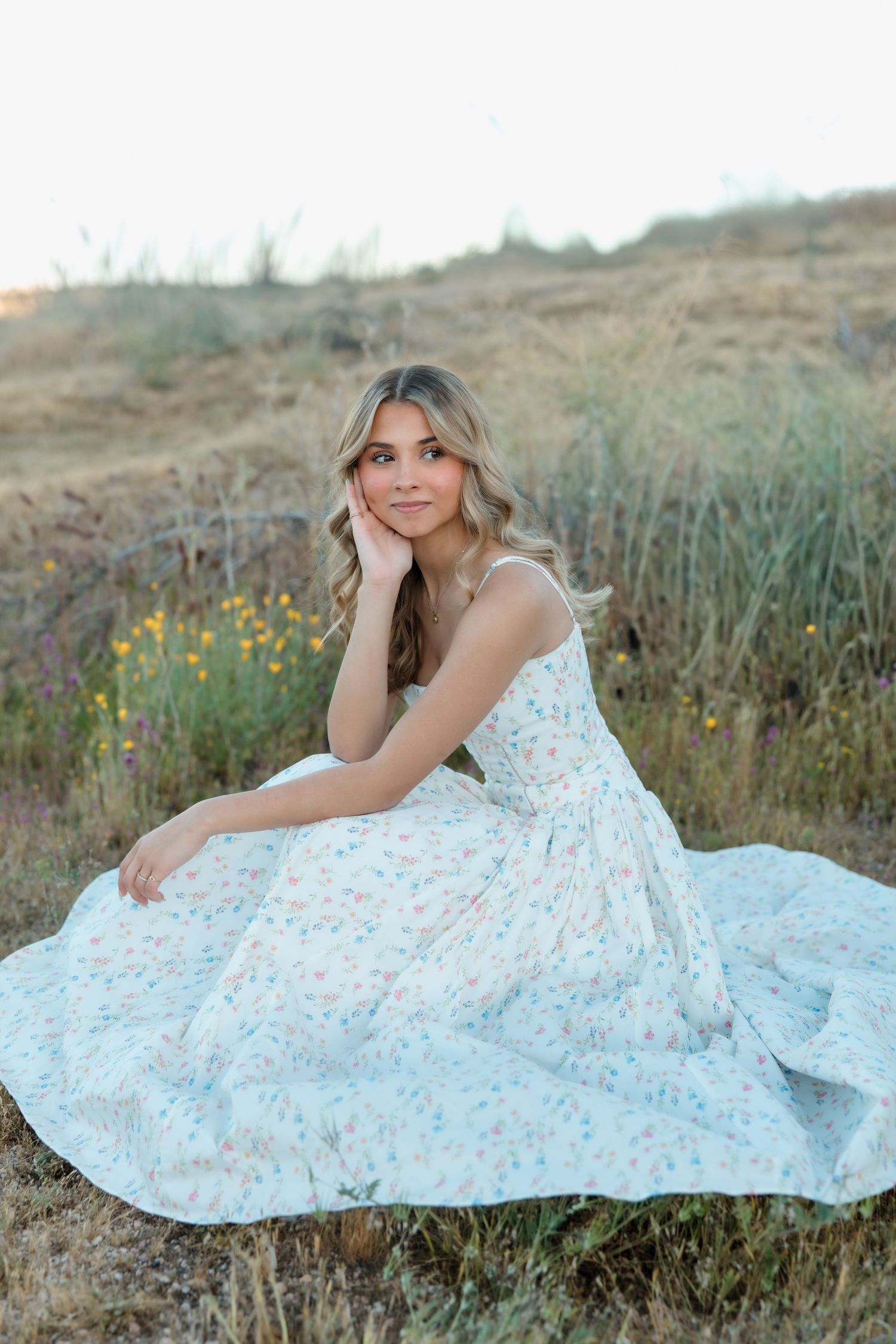 Young woman in floral maxi dress sitting gracefully in a golden field with wildflowers at sunset.