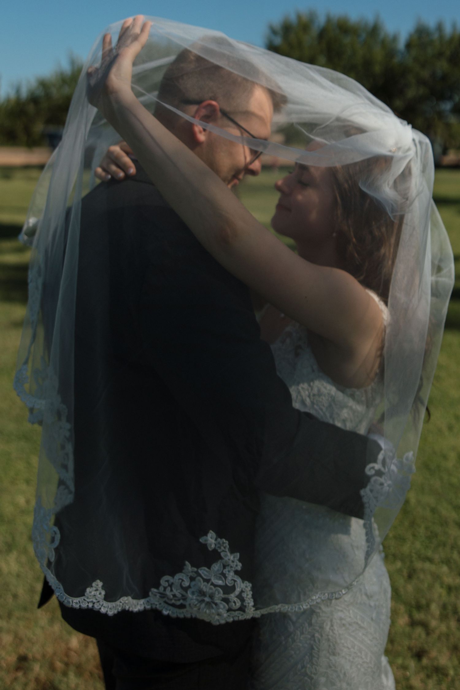 Bride and groom share romantic moment under lace wedding veil outdoors on sunny day.