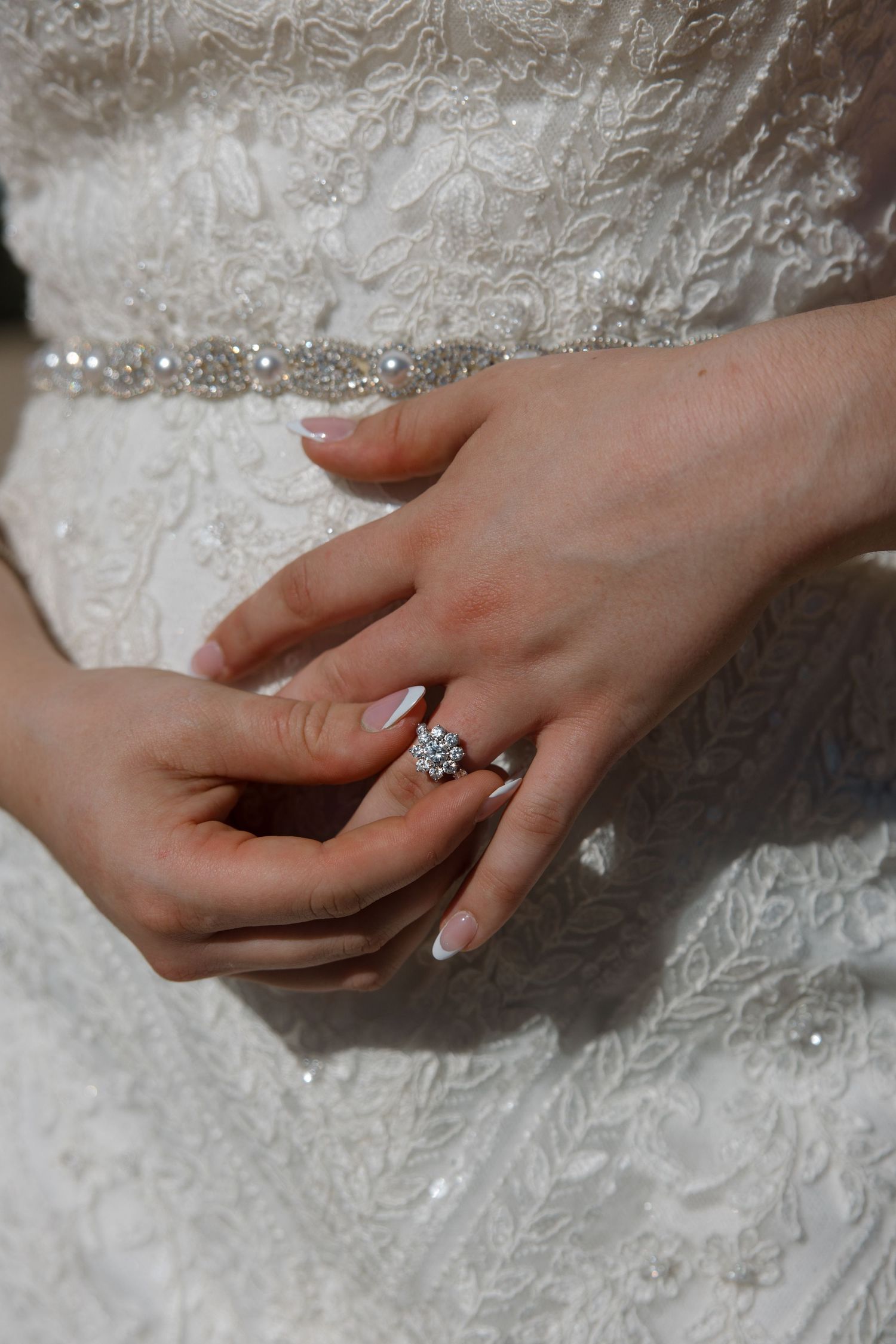 Close-up of a bride's hands showcasing a sparkling floral diamond engagement ring against her white lace wedding dress.