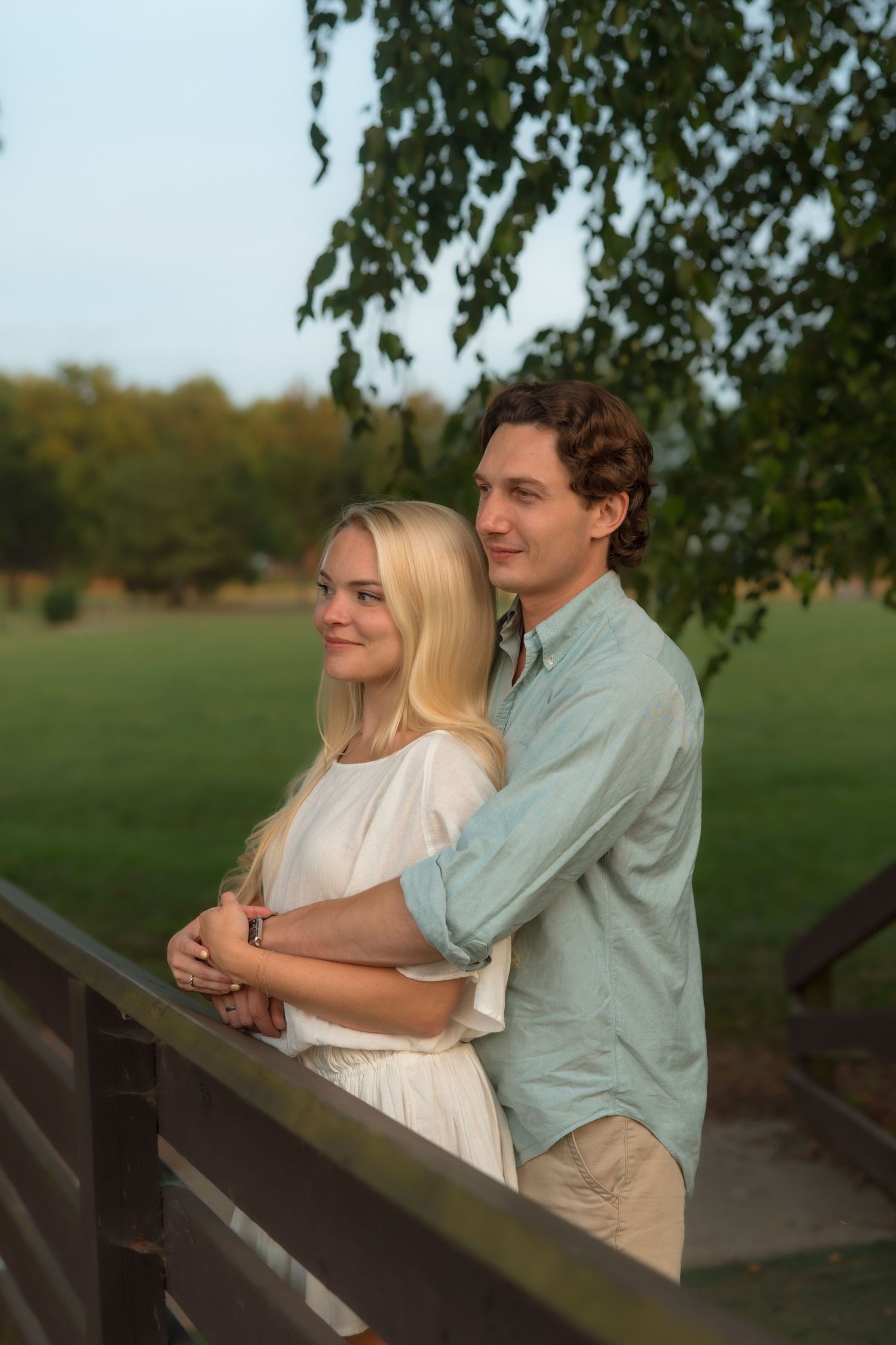 A smiling couple embraces on a wooden bridge, with the man hugging the woman from behind.