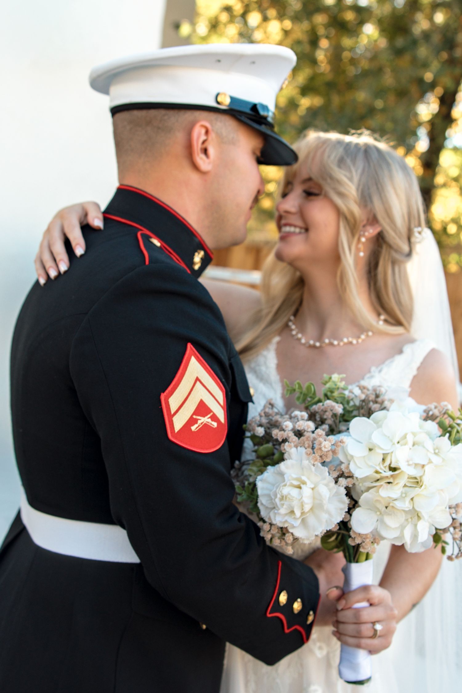 Marine groom in dress blues embraces smiling bride holding white hydrangea bouquet outdoors.