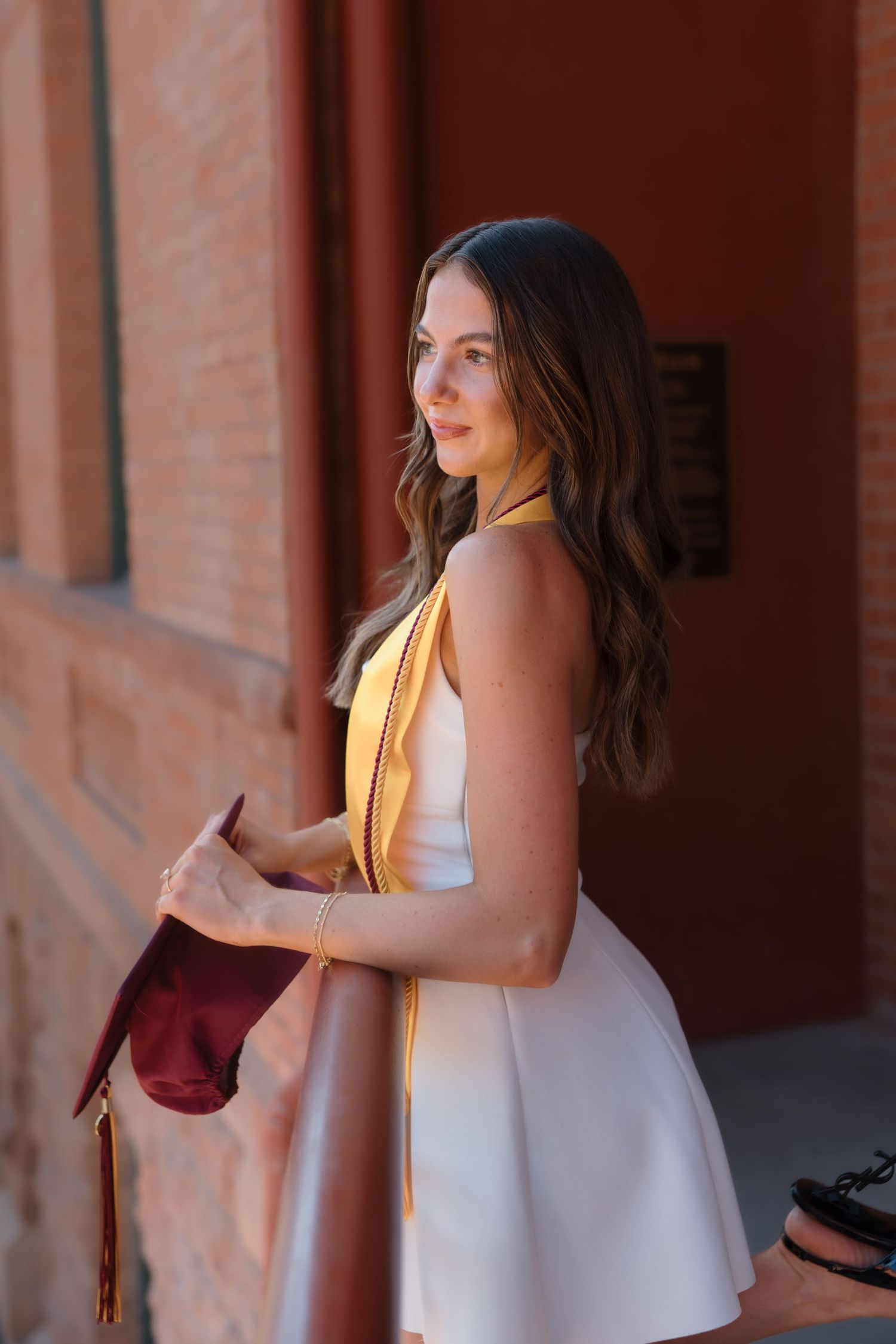 Brunette woman in white halter dress with gold ASU graduation stole holding maroon cap outdoors.