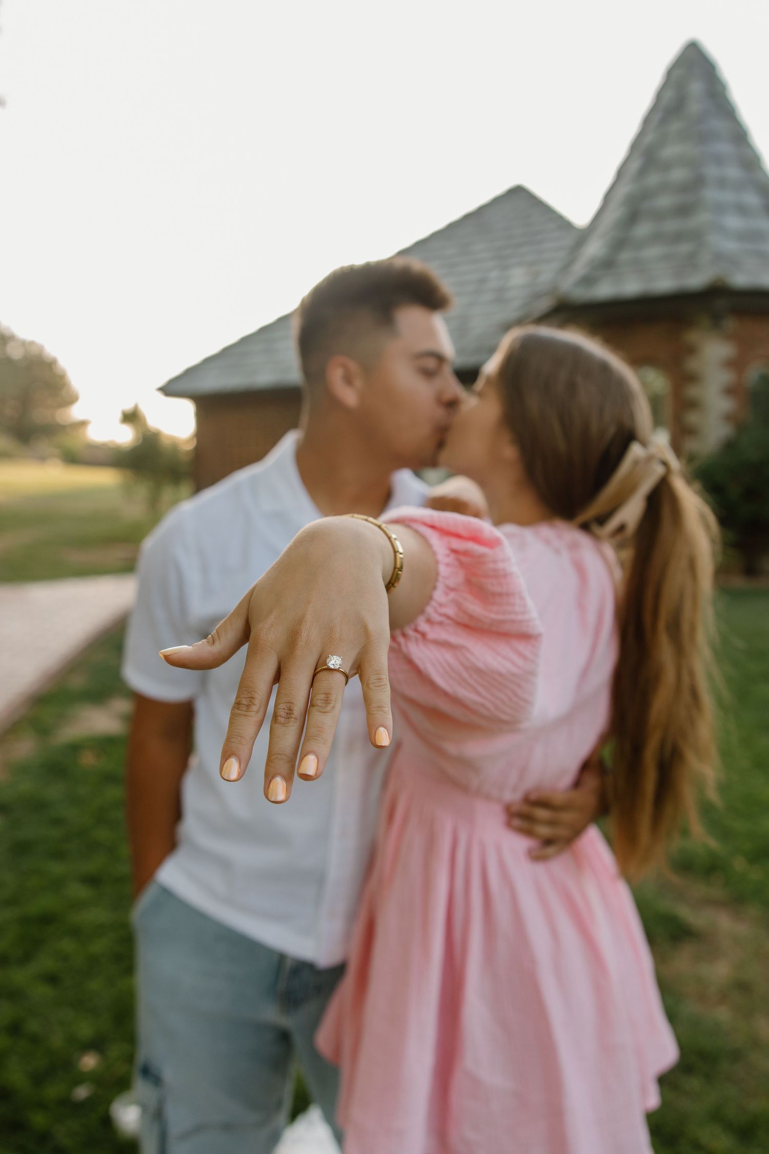 Couple kissing outdoors while showing off an engagement ring, wearing pink dress and white shirt near a rustic building.